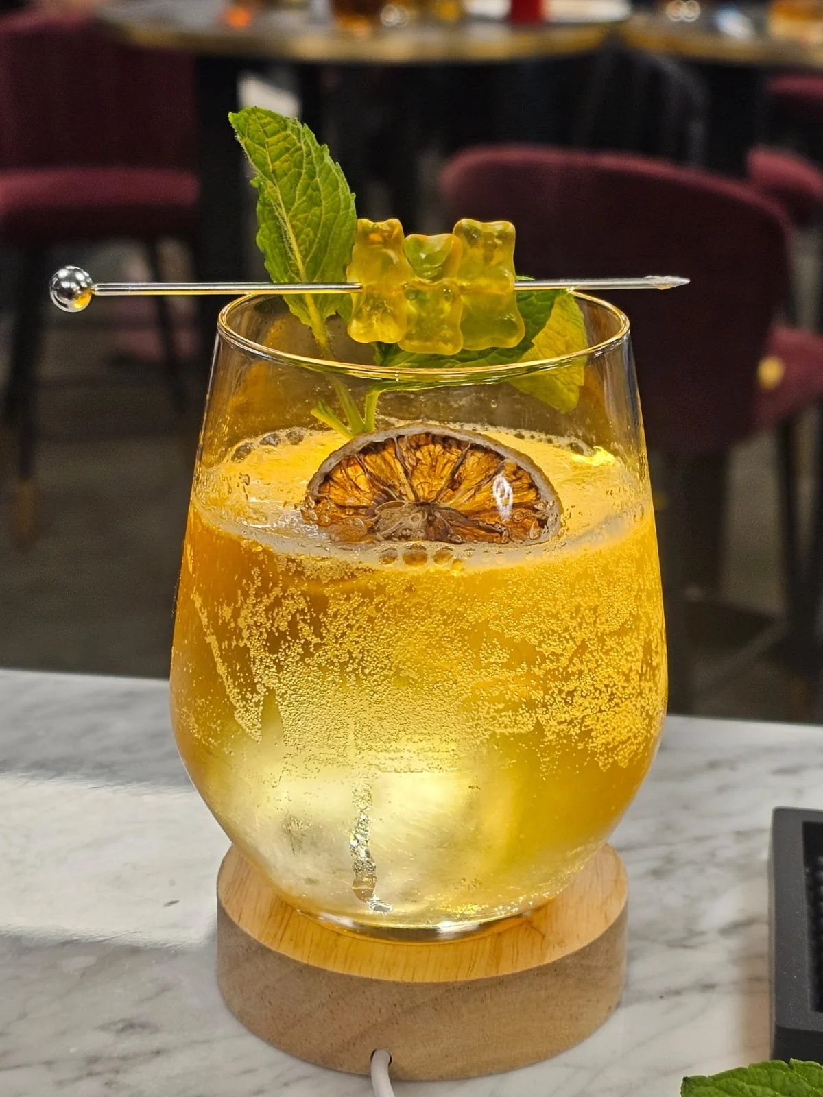 A glass of a yellowish carbonated beverage garnished with a slice of lime, a sprig of mint, and a gummy bear on a skewer, placed on a wooden coaster on a marble surface.