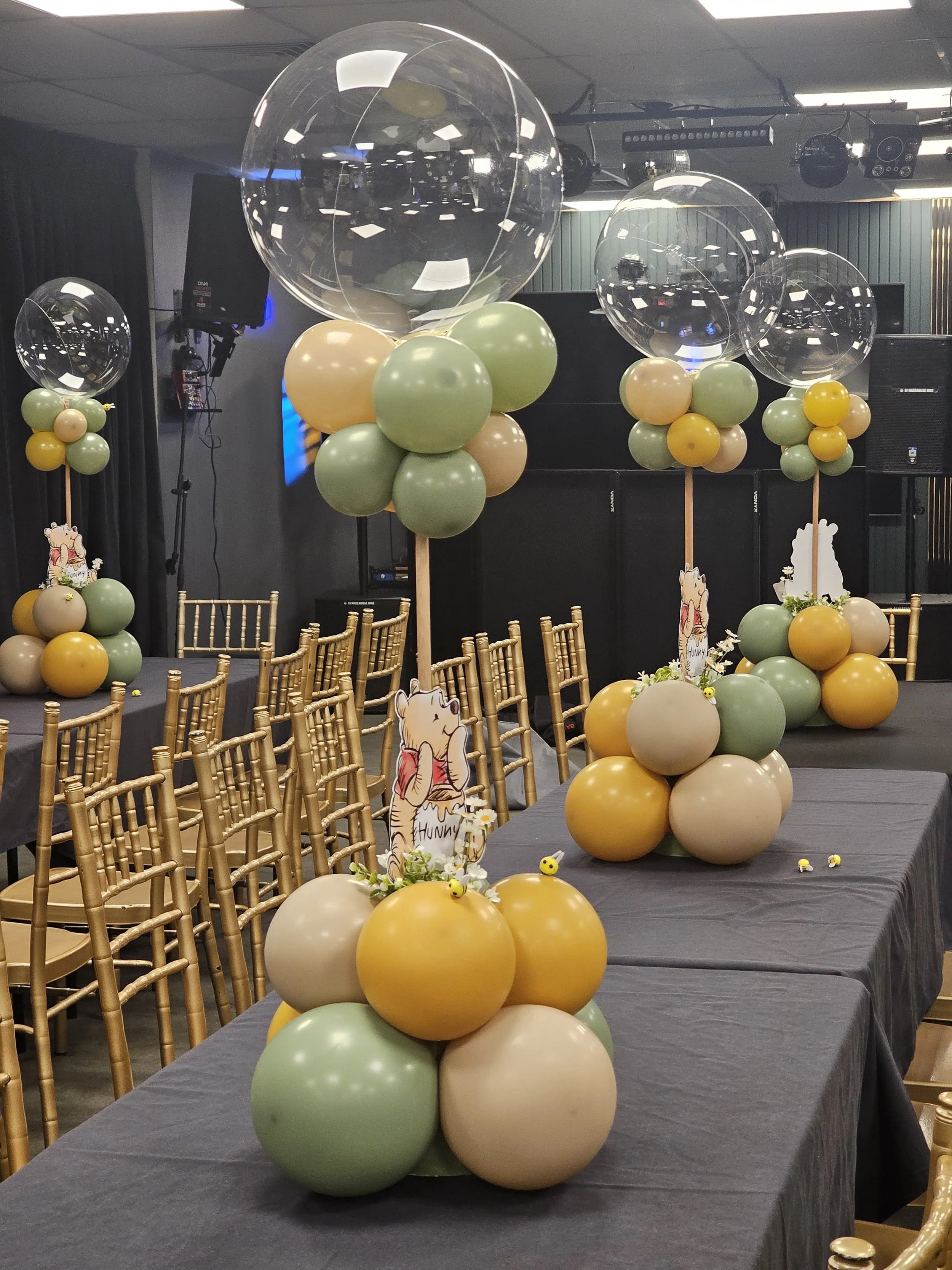 Balloon centerpieces with large clear balloons and smaller balloons in shades of green, yellow, and beige, arranged on tables with gold chairs, in a decorated event space.
