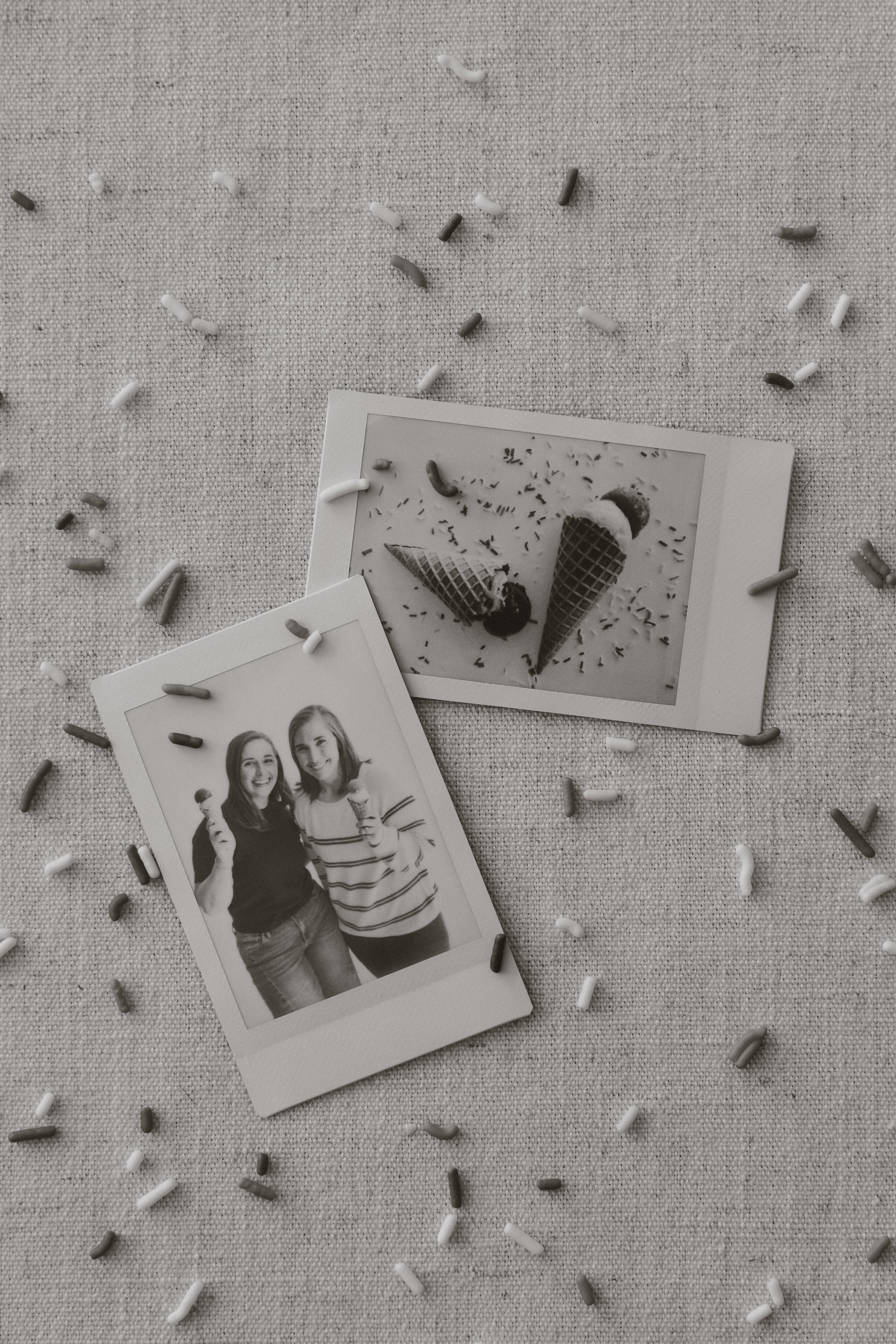 Black and white photograph of two young women smiling, holding ice cream cones, standing close together, with sprinkles scattered on the surface below. A second photo of a similar scene is partially visible.