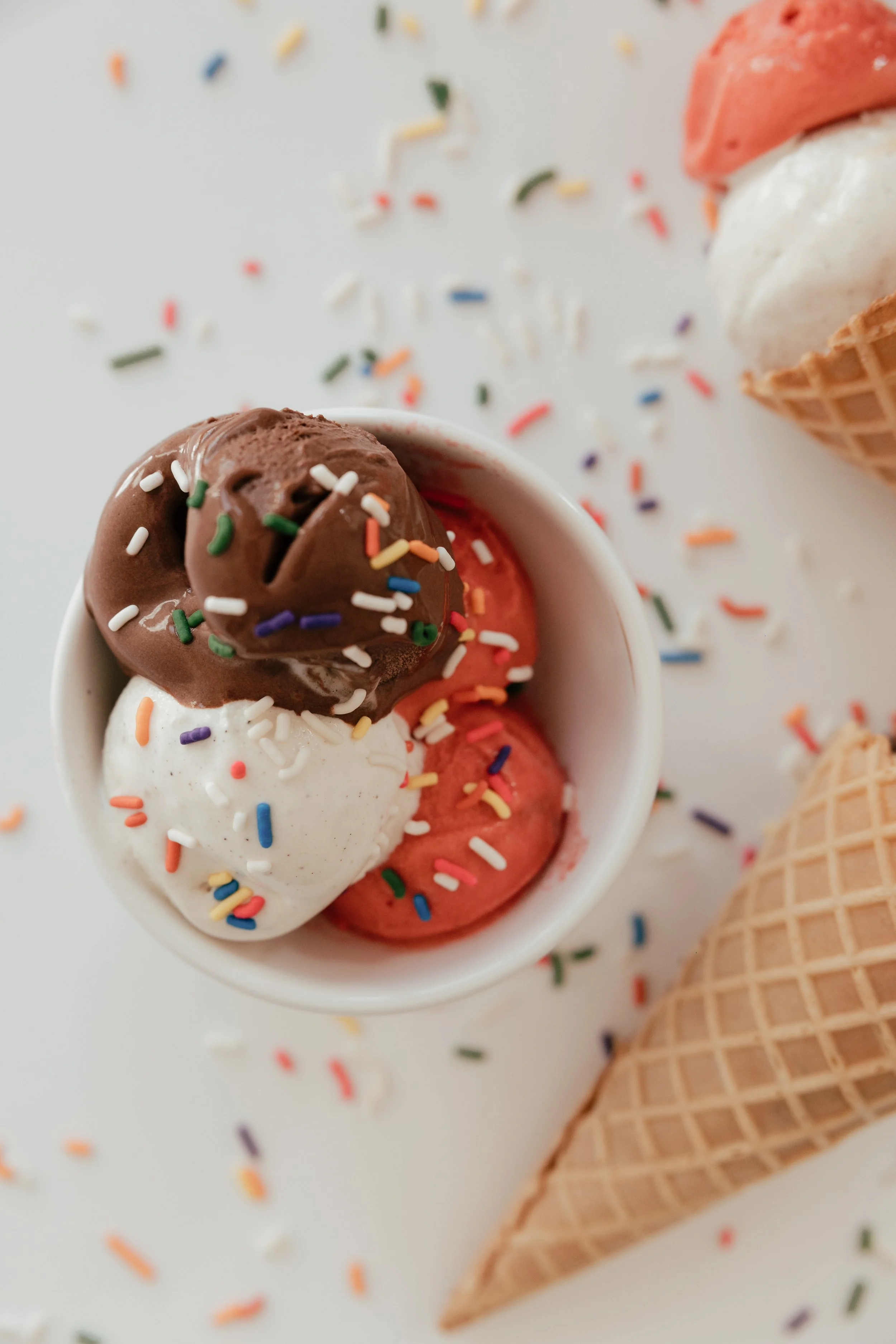 A cup of ice cream scoops with rainbow sprinkles, topped with a chocolate scoop, alongside an ice cream cone with two scoops, one white and one pink, surrounded by scattered rainbow sprinkles.