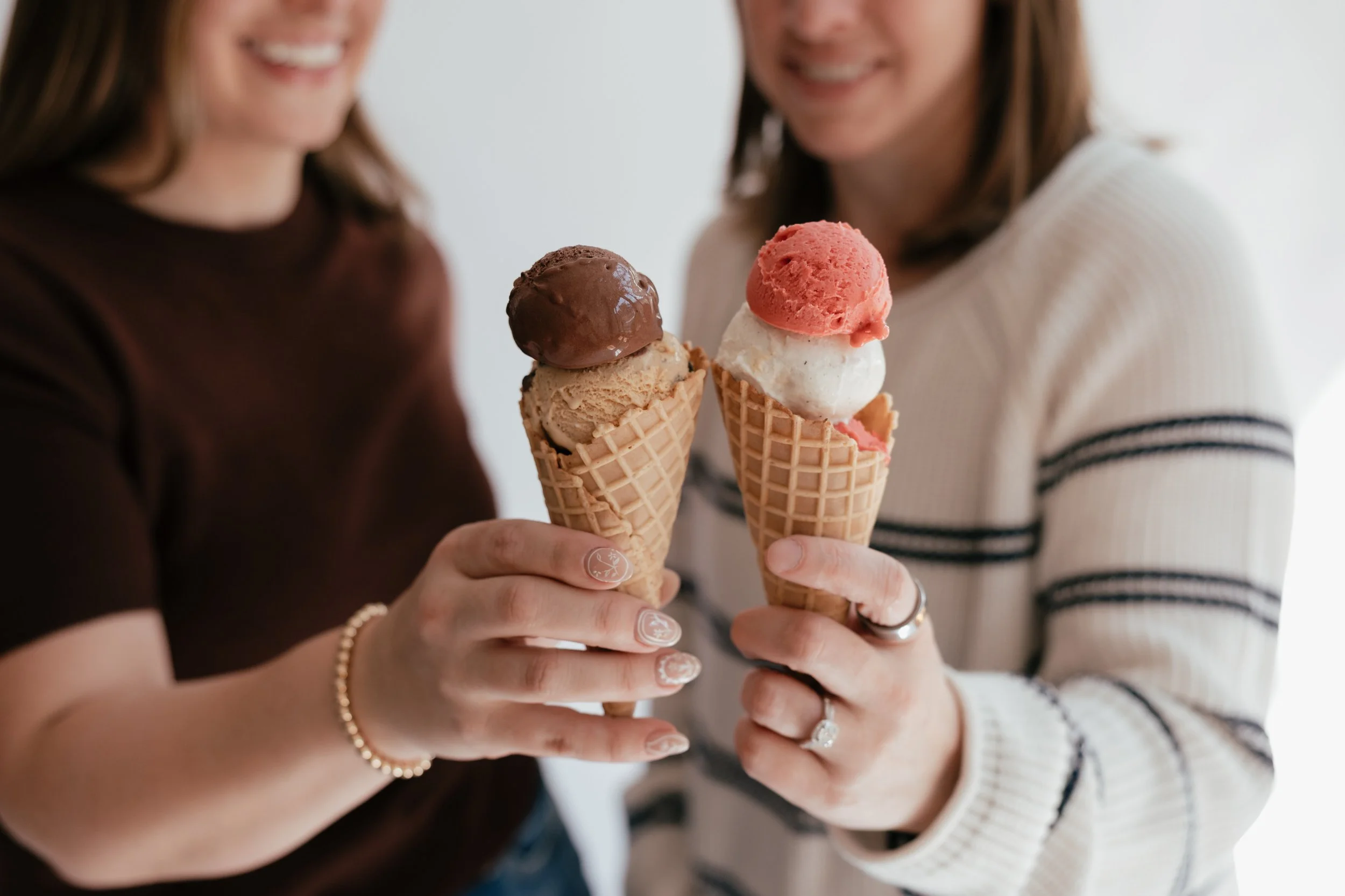 Two women holding ice cream cones topped with three scoops each, smiling at each other.