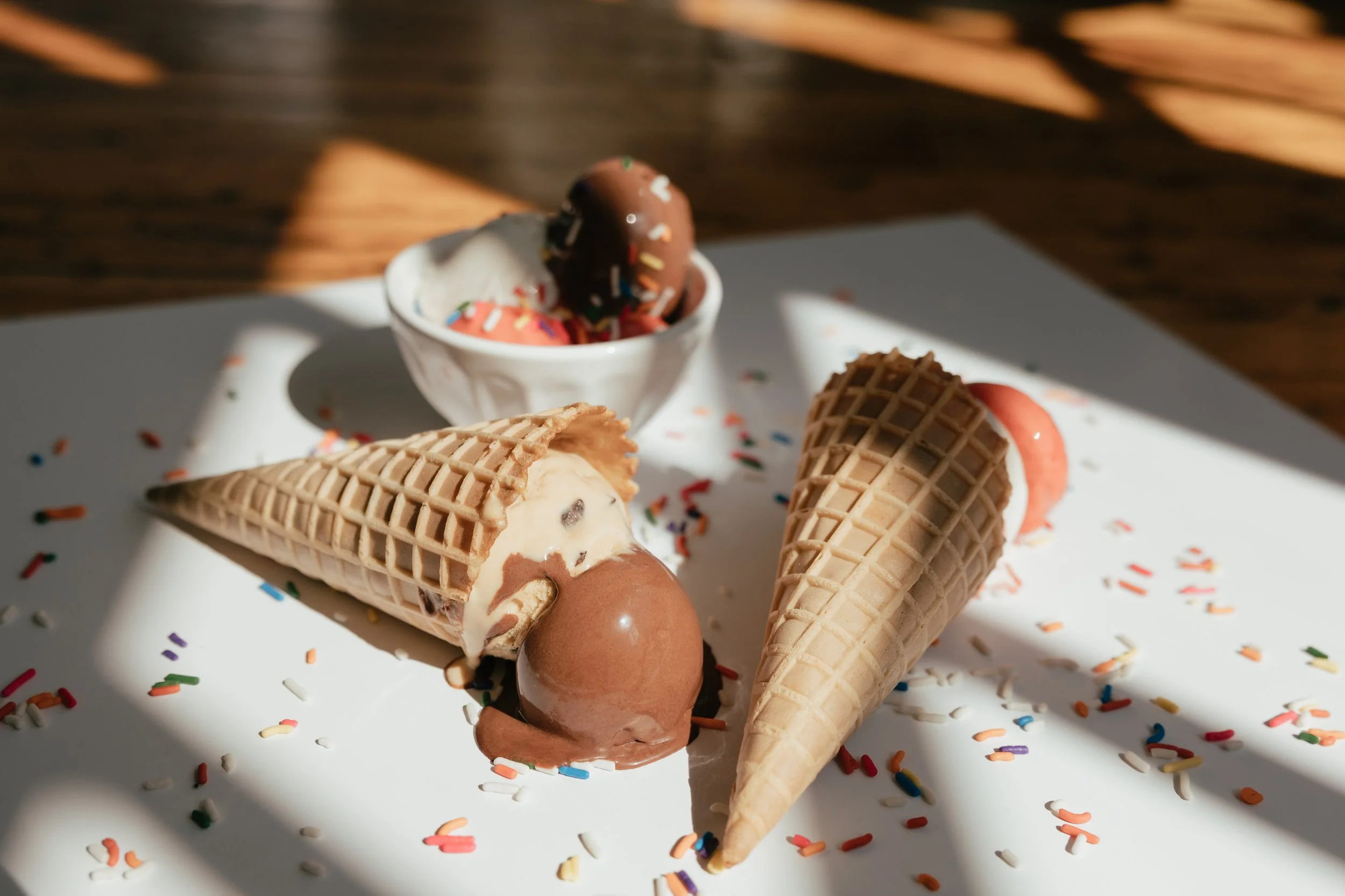 Three ice cream cones and a small bowl of ice cream with colorful sprinkles on a white plate, surrounded by scattered confetti on a white surface with a wooden background.