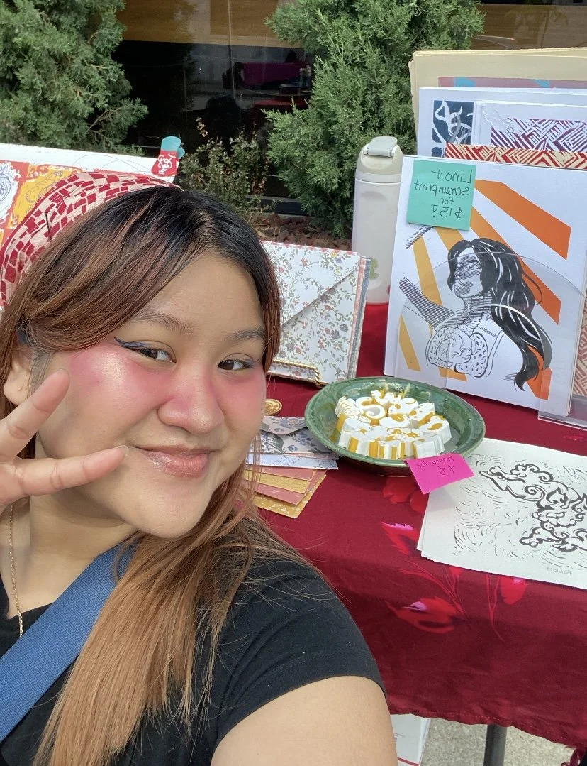 A young woman taking a selfie at a table with artwork and crafts. She is smiling and making a peace sign. The table has drawings, a earthenware plate with white and yellow decorative items, and a pink sticky note. Behind her, there are two green bushes and a window.