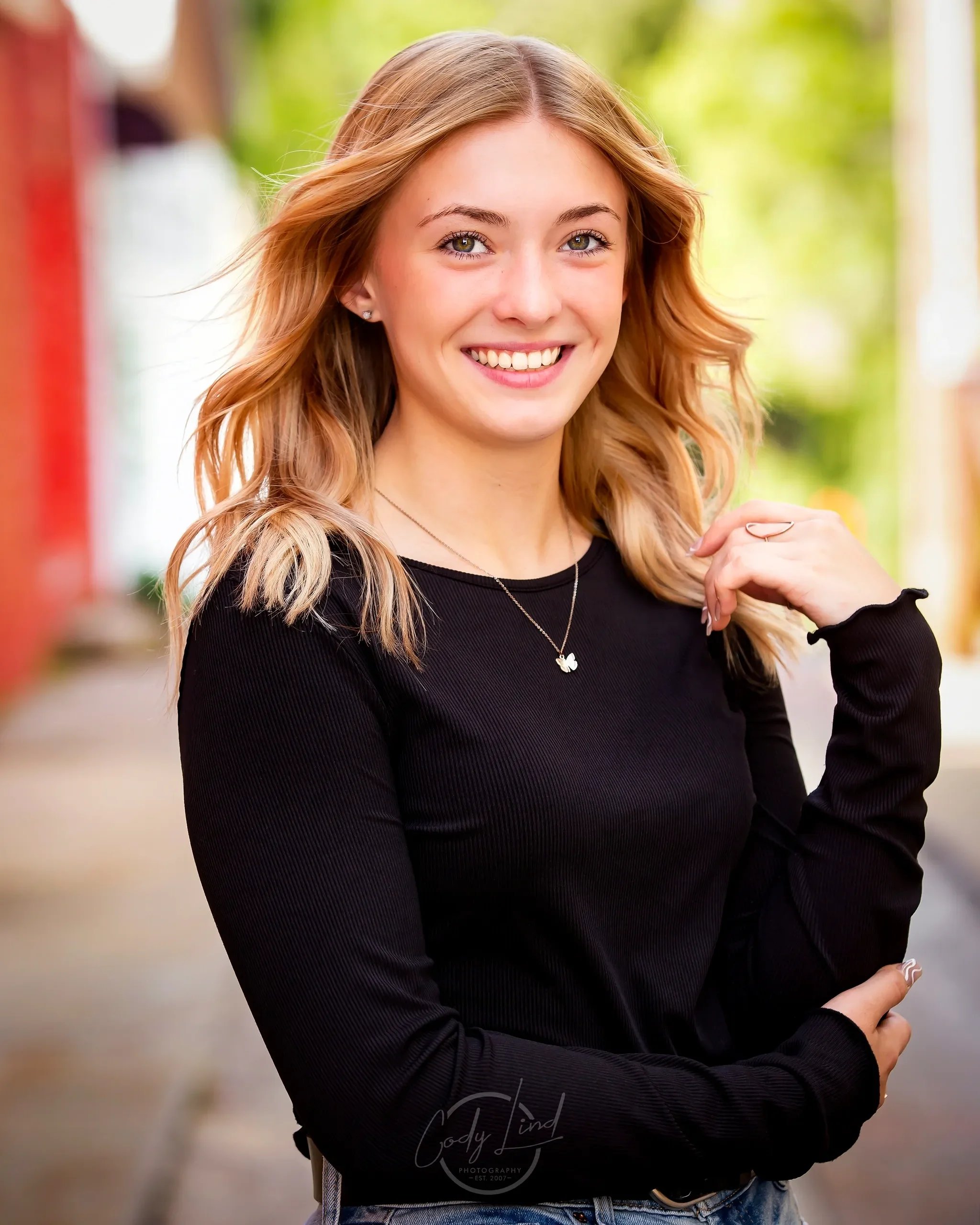A young woman with blonde, wavy hair and green eyes smiling outdoors, wearing a black long-sleeve top and a silver necklace with a butterfly pendant.