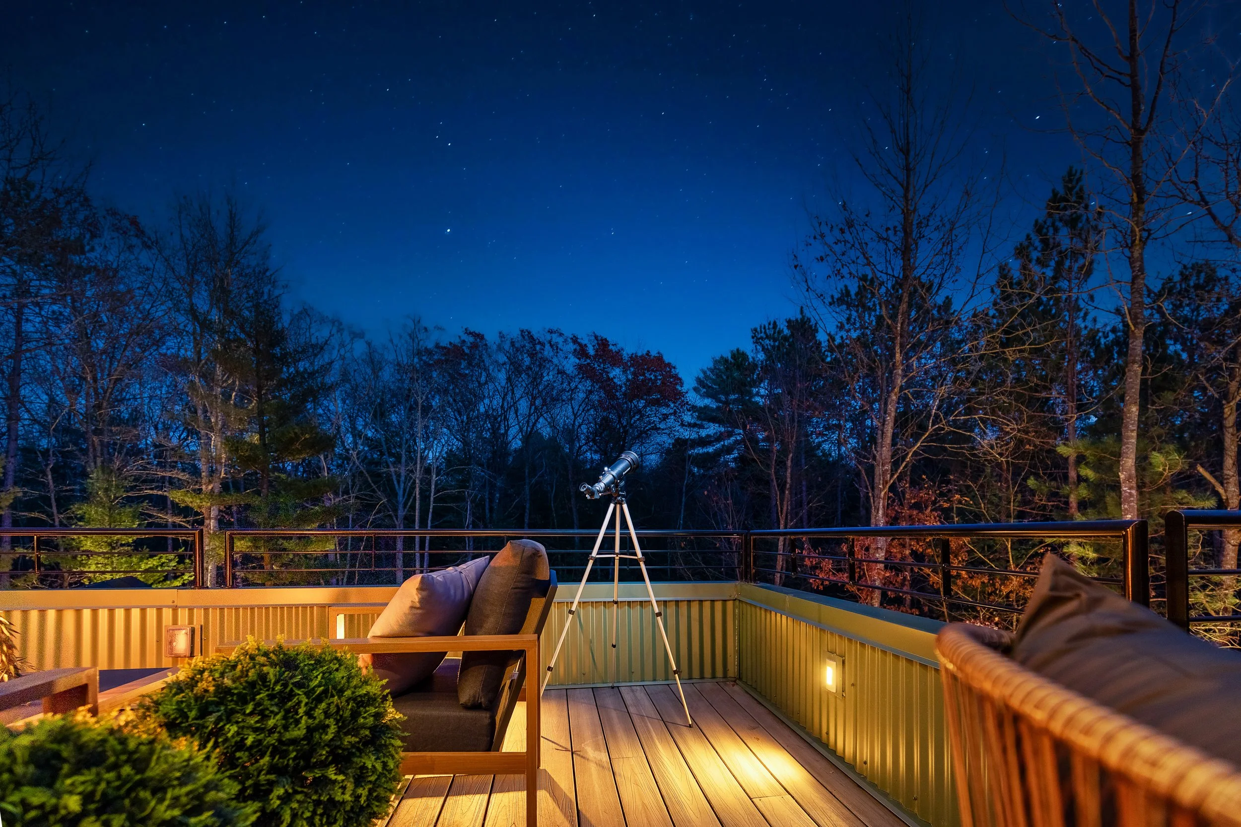 Nighttime outdoor patio with telescope, lounge chairs, and string lights, overlooking a wooded area with clear starry sky.
