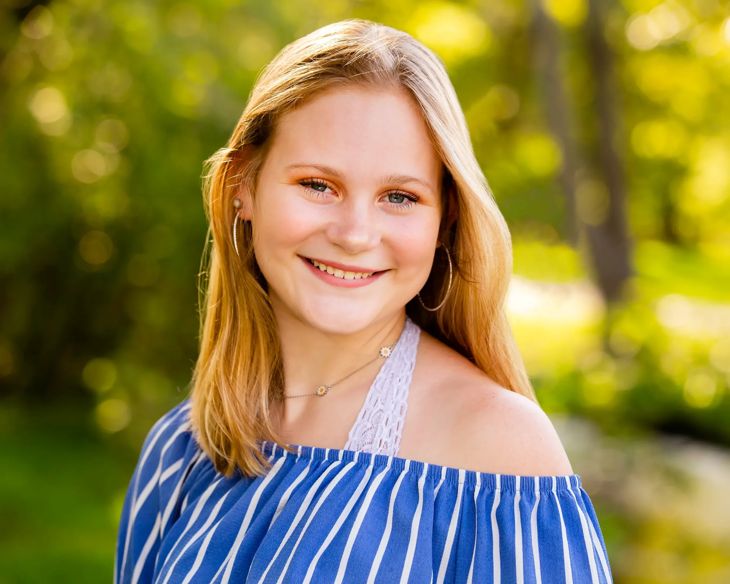 A young woman with blonde hair and blue eyes smiling outdoors with a blurred green background.
