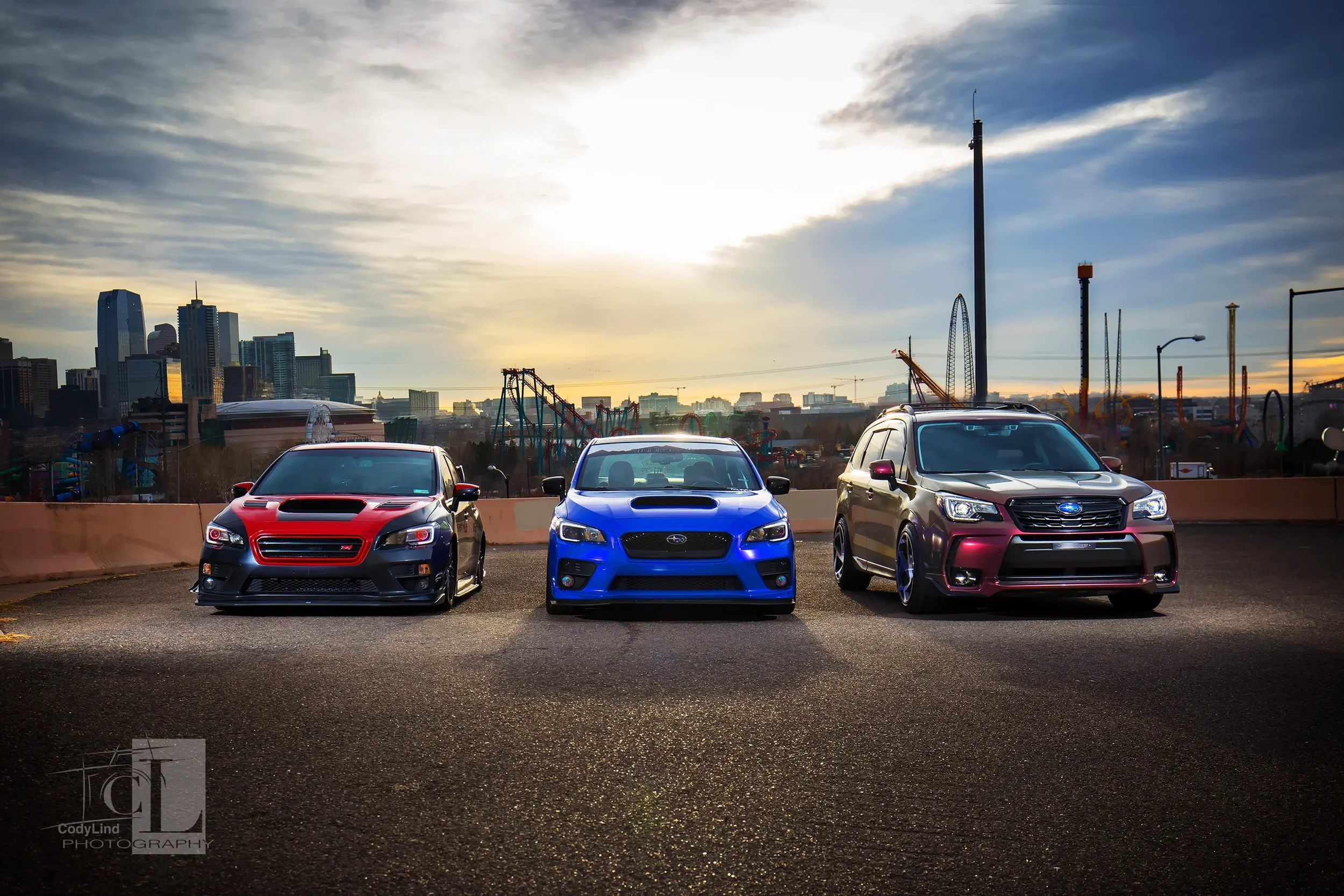 Three cars parked side by side on a rooftop parking lot with a city skyline and amusement park rides in the background during sunset.