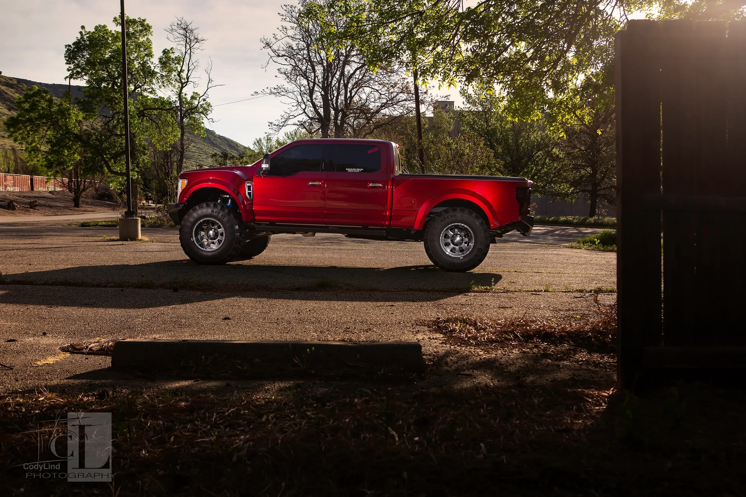 A red pickup truck parked in an empty parking lot during late afternoon or early evening with trees and mountains in the background.