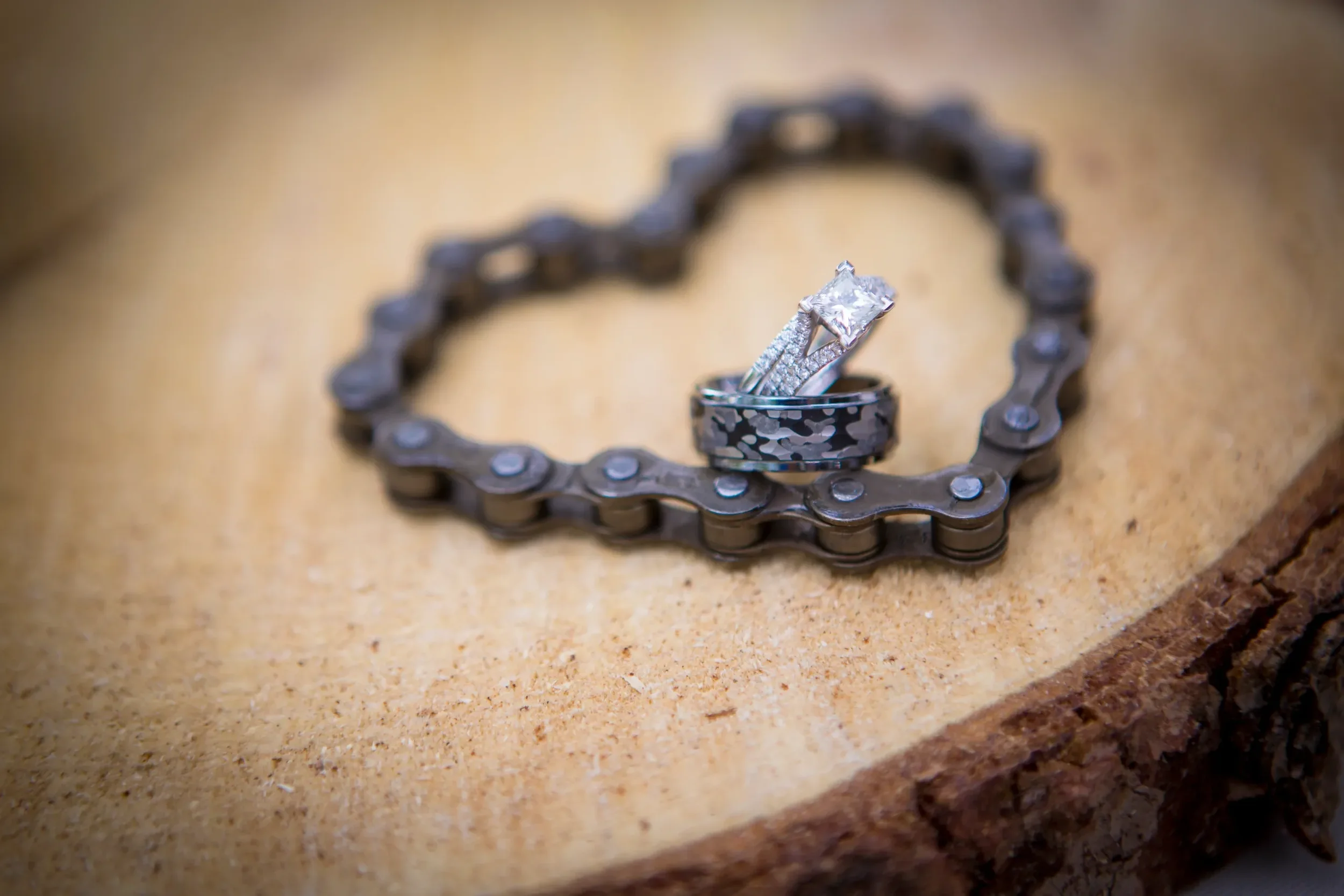 A diamond engagement ring and wedding band placed on top of a metal bicycle chain, which is arranged in a heart shape on a wooden surface.