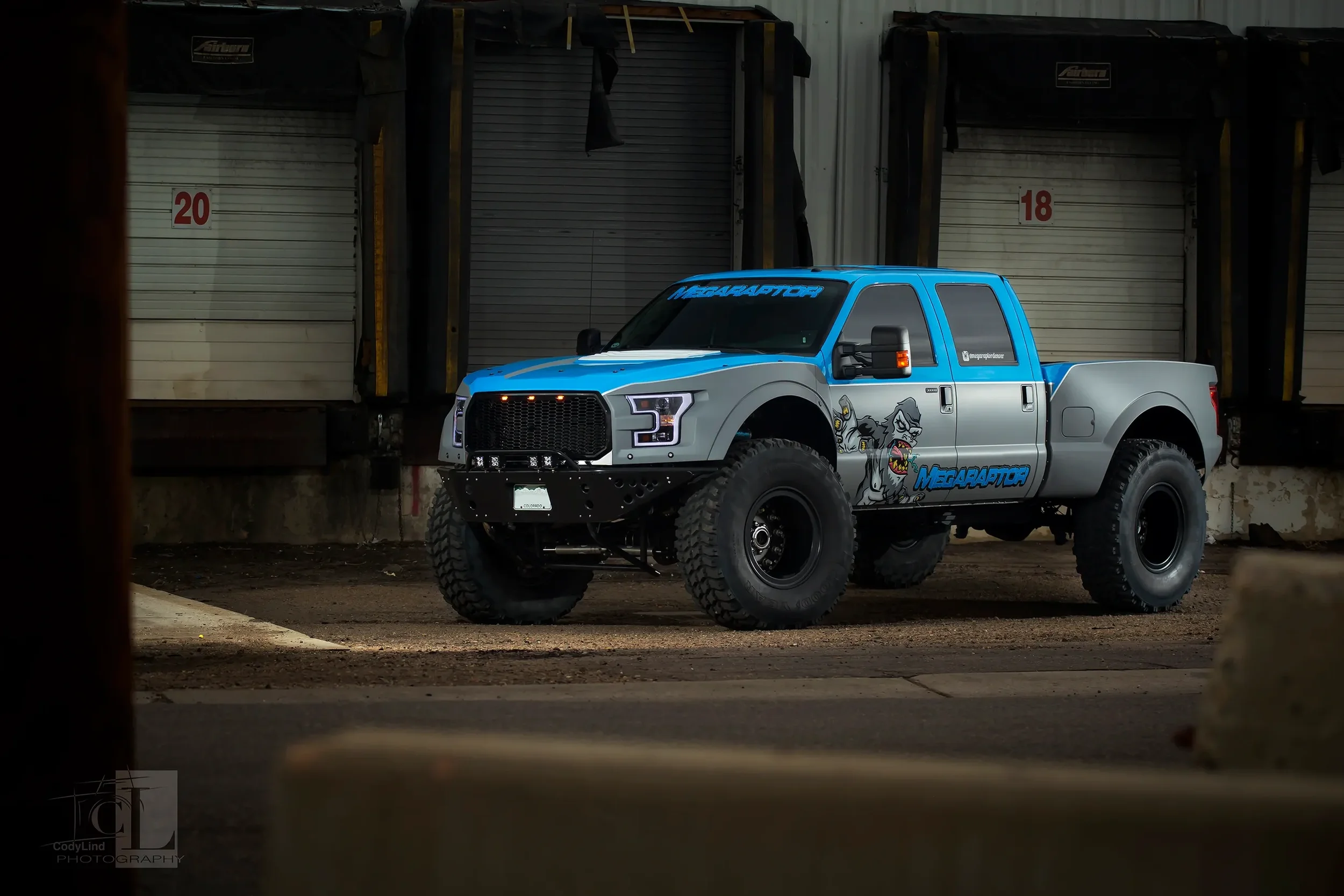 A custom off-road truck painted with blue and gray colors, featuring the 'Megaraptor' logo and cartoon dinosaur graphics, parked on a dirt lot in front of closed storage unit doors.