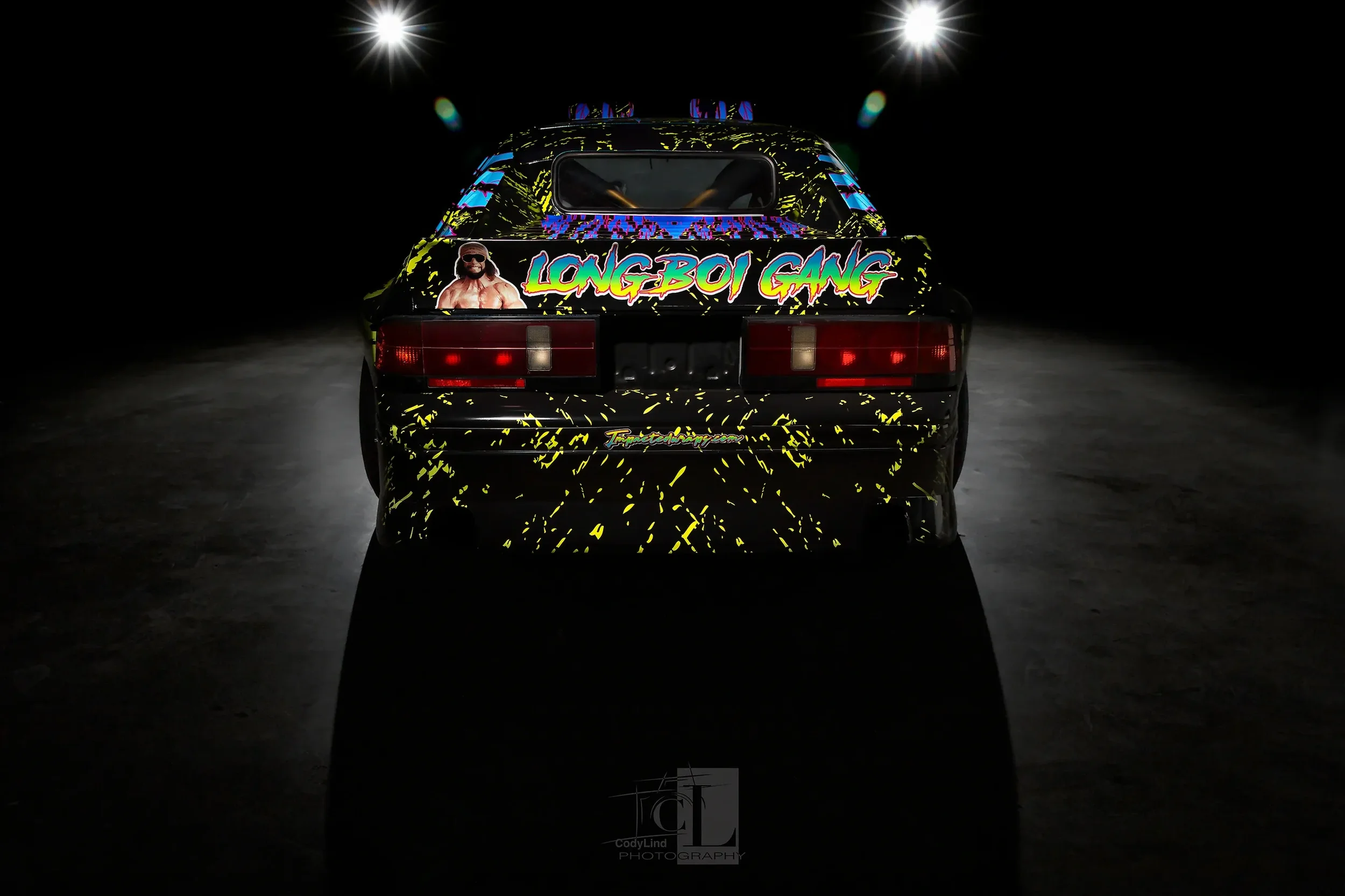 Rear view of a black sports car with colorful neon-style graphics, including the words "LONGBOI GANG" and an image of a shirtless man with sunglasses, set against a dark background with two bright light sources overhead.