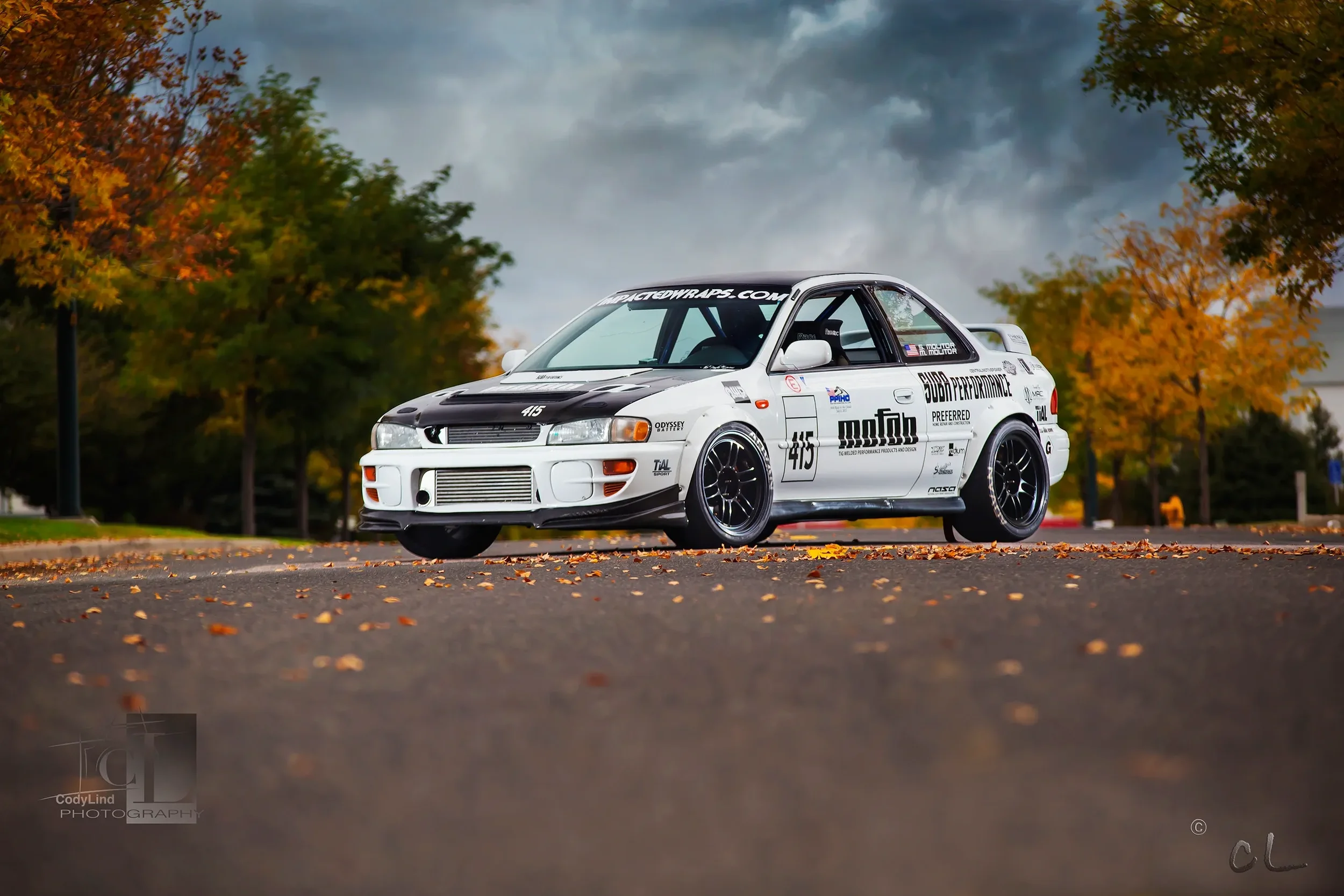 A white rally car with black accents and various sponsor decals parked on an asphalt road with fallen leaves. Autumn trees with colorful foliage are in the background under a cloudy sky.