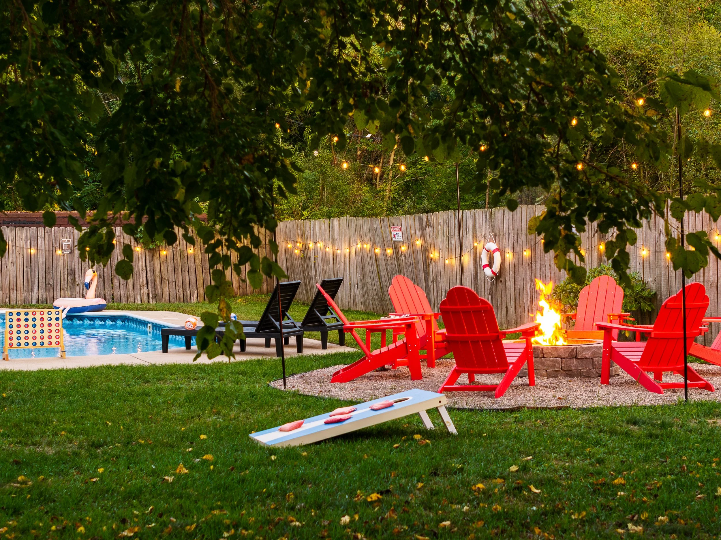 Backyard scene at dusk with a swimming pool, black and red Adirondack chairs around a fire pit, string lights overhead, and a wooden fence with a life preserver and a sign.