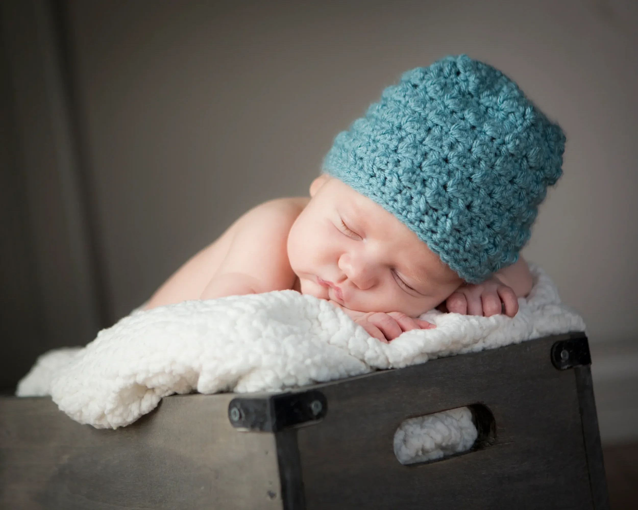 A sleeping baby with a blue knitted hat, resting on a soft blanket in a wooden box.