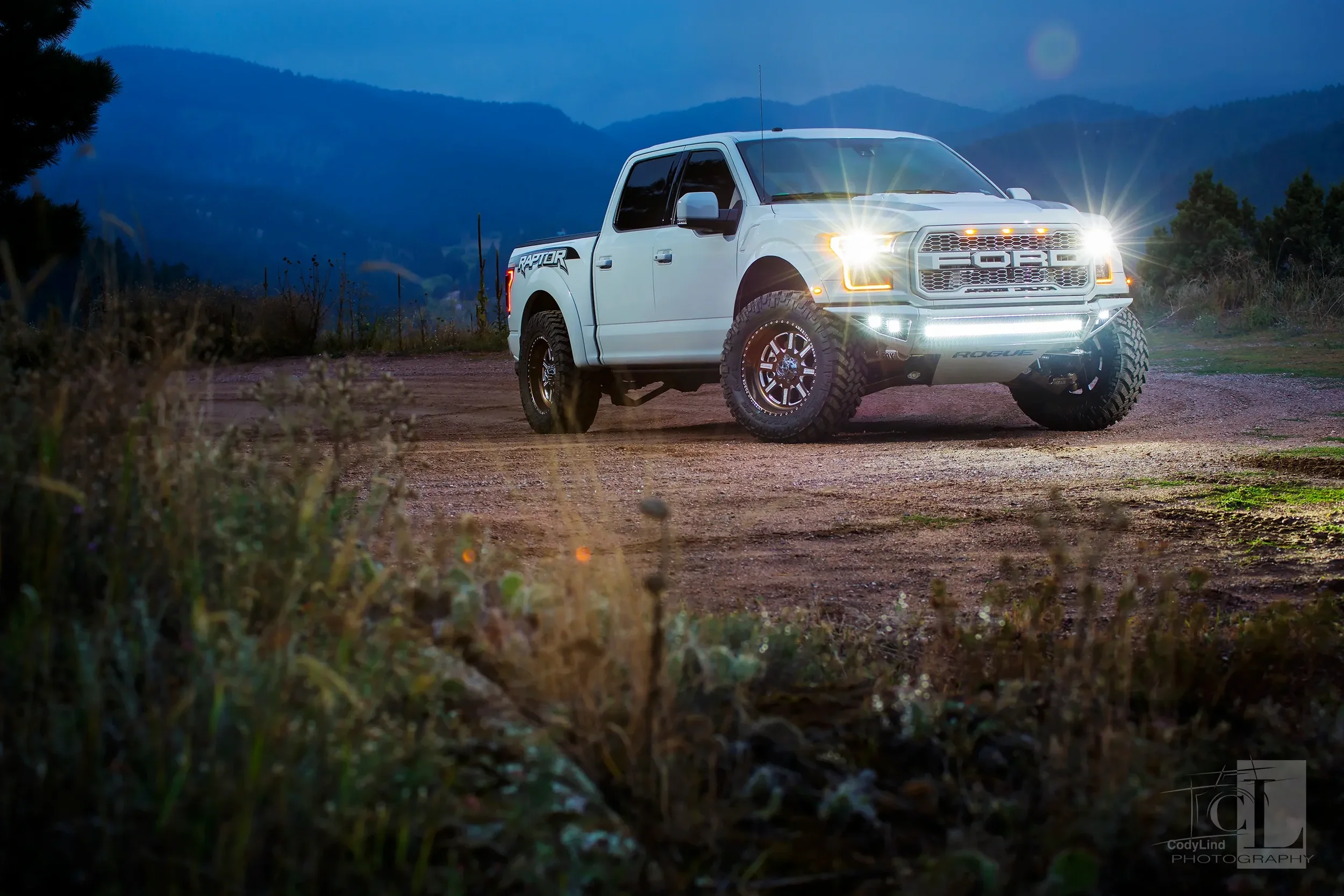 White off-road pickup truck with bright headlights parked on dirt trail in mountains at dusk.