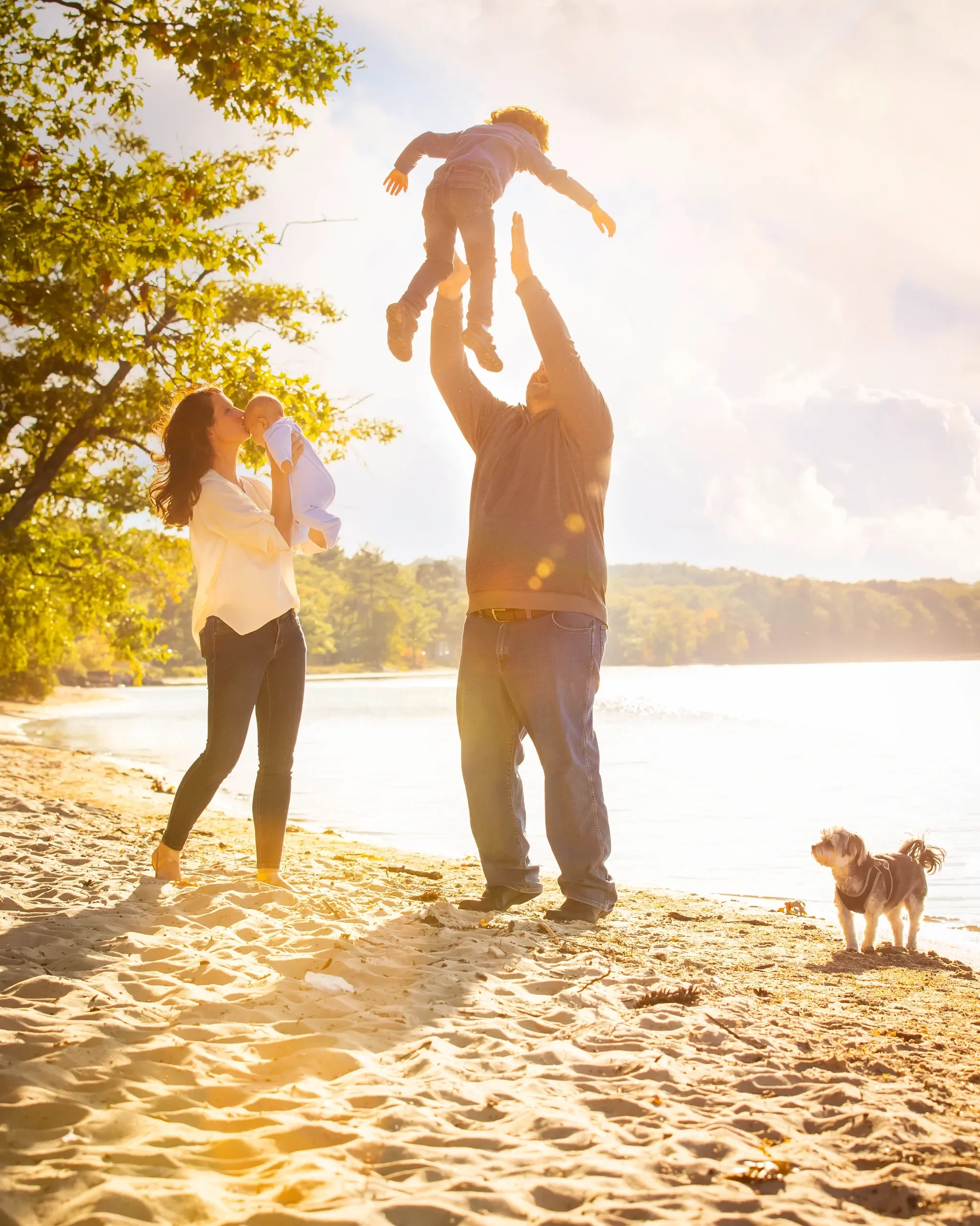 Family on the beach with a man tossing a child in the air, a woman holding a baby, and a dog standing nearby, during sunset.