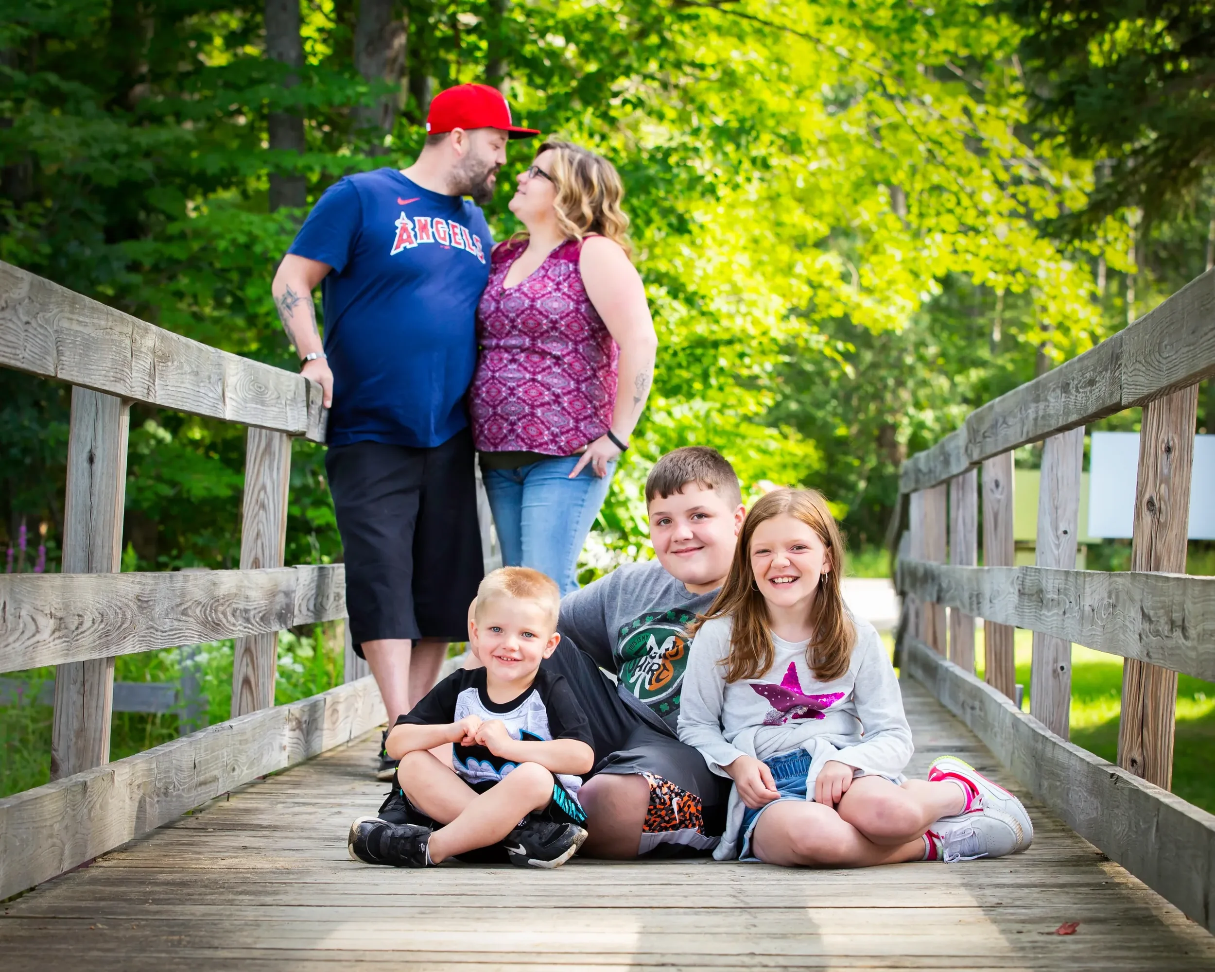 A family of five on a wooden bridge in a green, wooded park. Two adults stand together at one end, gazing at each other, while three children sit or lie on the bridge in the foreground, smiling.