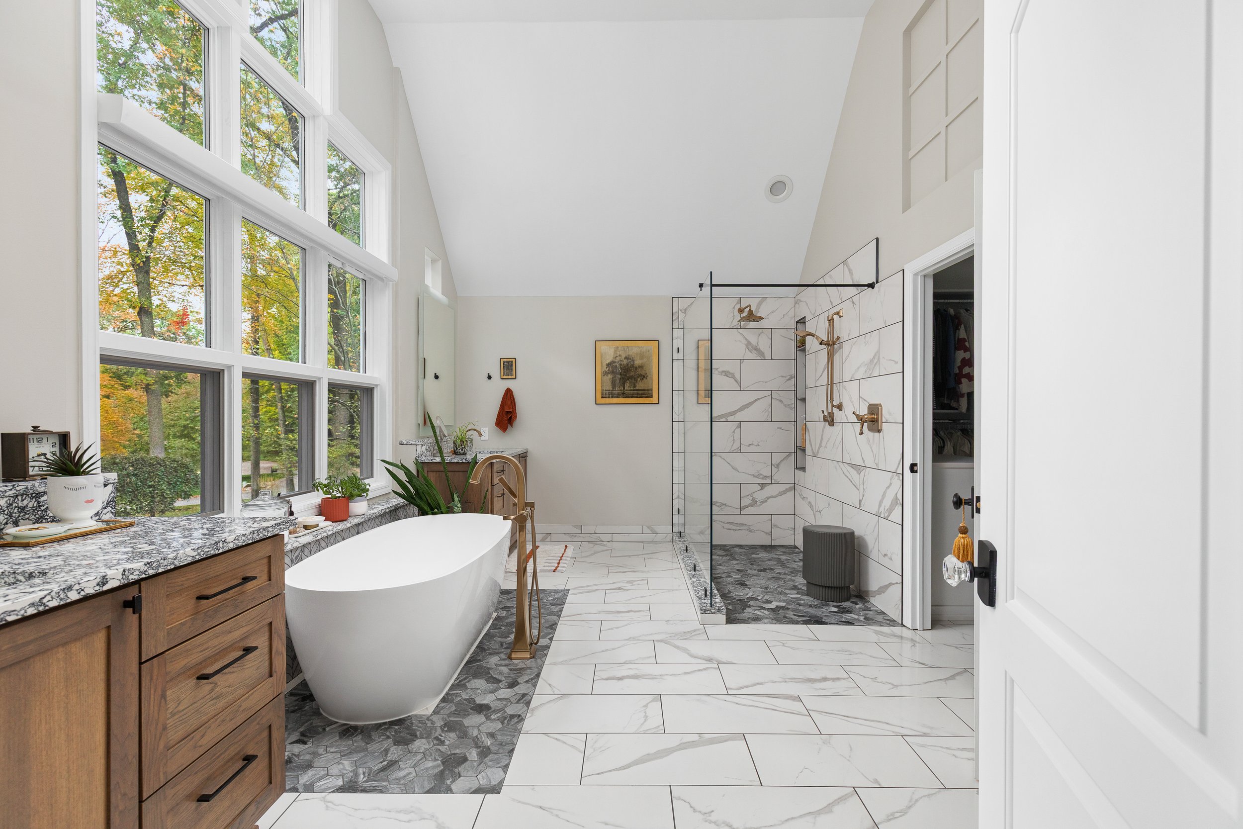 Modern bathroom with white marble floors, large windows, a freestanding bathtub, plants, a vanity with a granite countertop, and a walk-in shower with marble walls and brass fixtures.