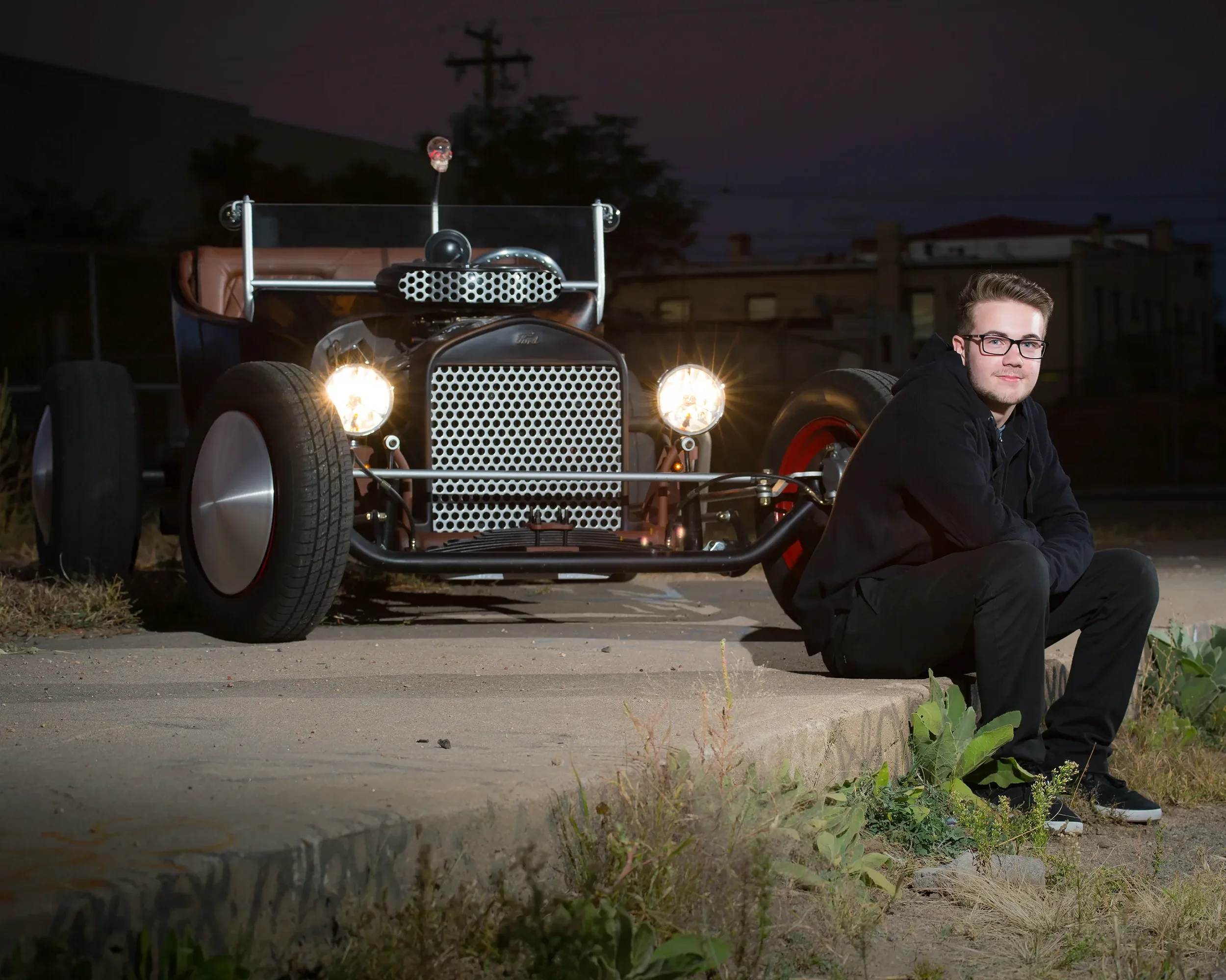 A young man with glasses and a black hoodie sits on a curb at dusk next to a vintage hot rod with its headlights on, parked on a sidewalk with buildings in the background.