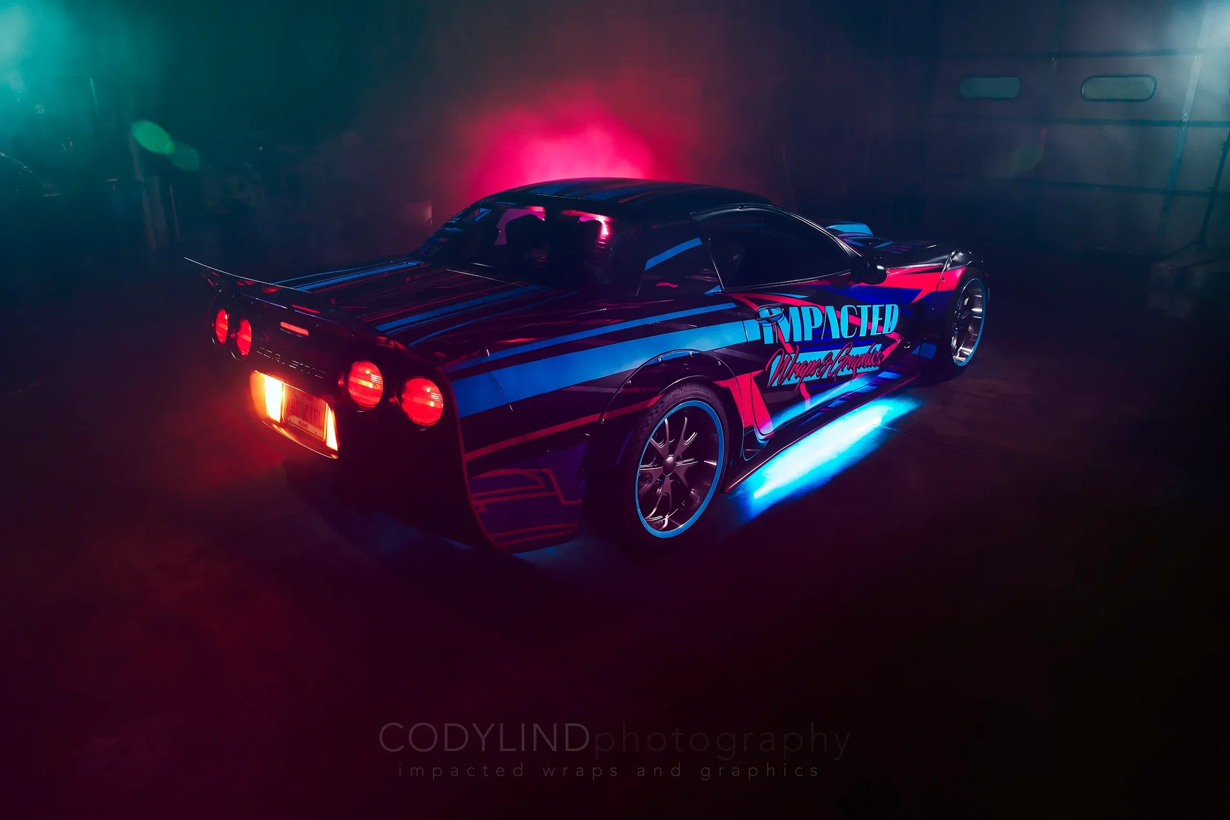 A sports car with a black body featuring pink and blue graphics and the words "Impacted Wraps & Graphics," illuminated by underglow lights, parked in a dimly lit garage with colorful lighting effects.