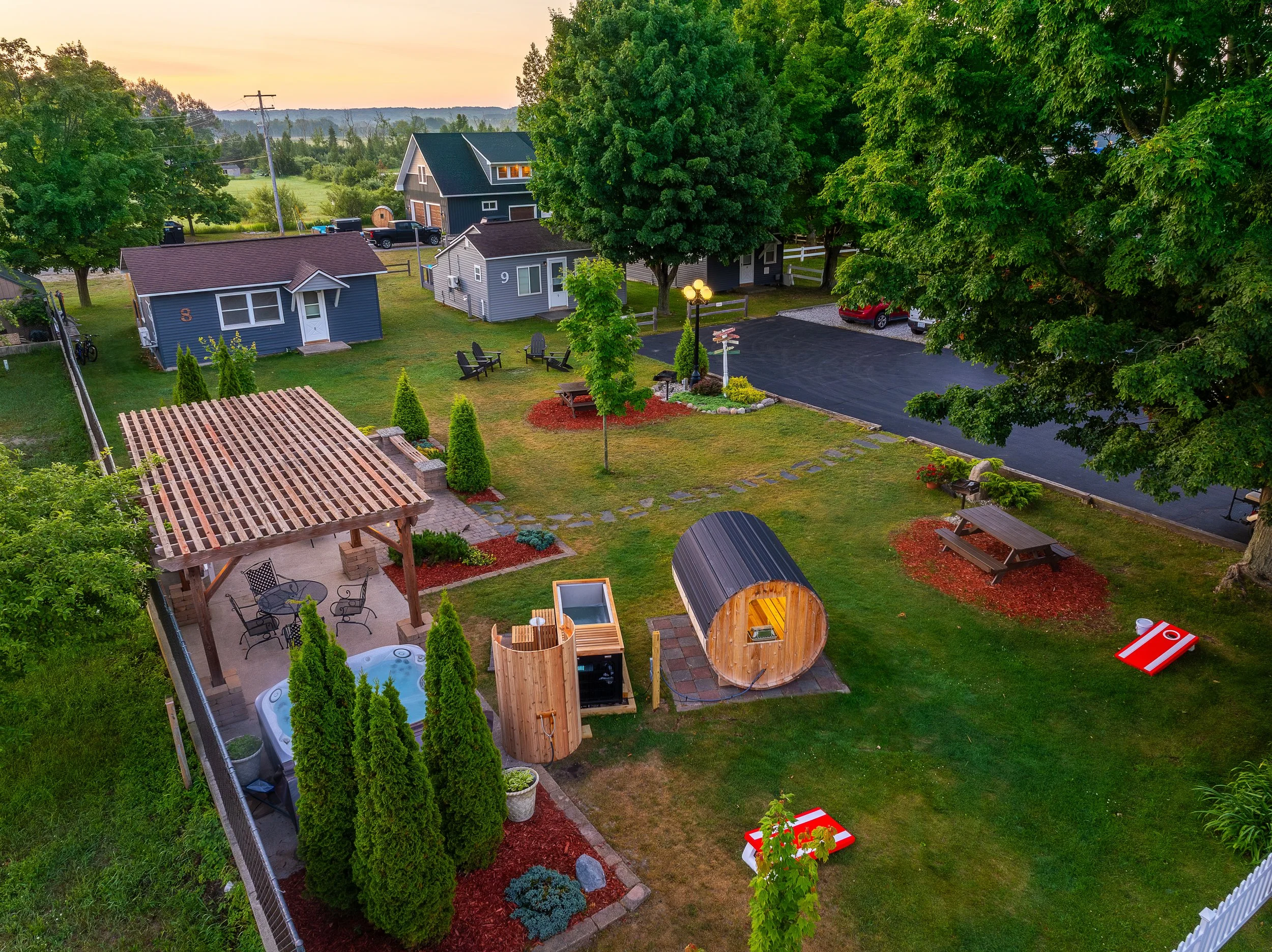 A backyard with a wooden pergola patio, hot tub, small wooden sauna, two cornhole games, picnic tables, a fire pit, and lush green trees and grass, with a parking lot and houses in the background at sunset.