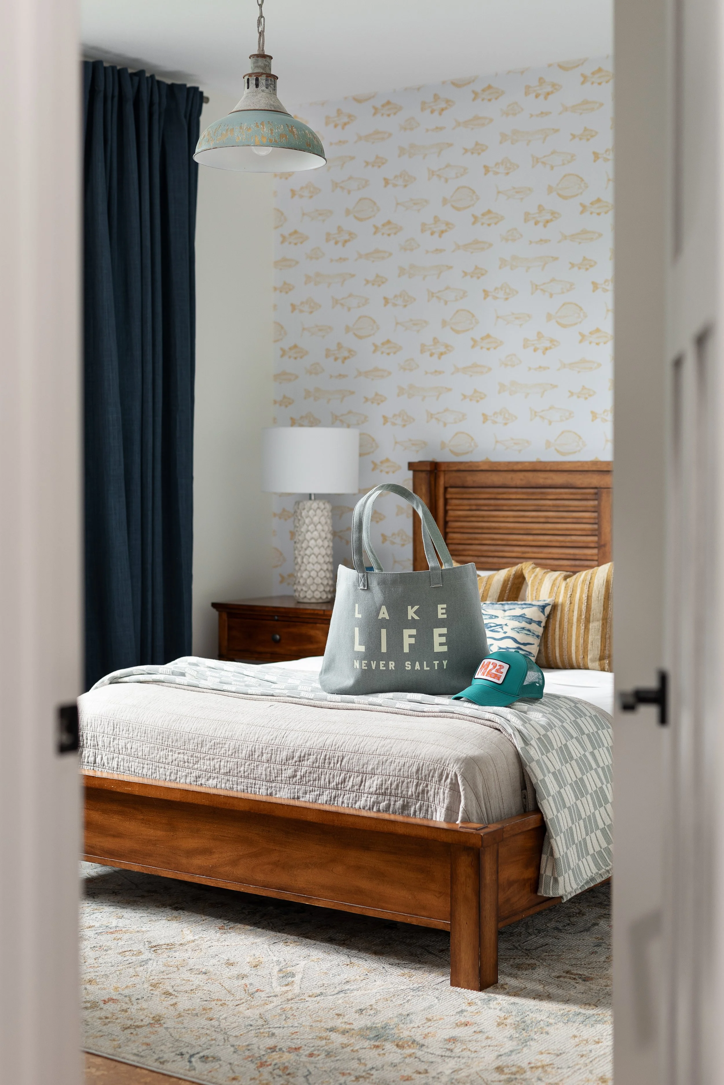 A bedroom with a wooden bed frame, striped and patterned pillows, a bedside table with a textured white lamp, navy curtains, a fish-themed wallpaper accent wall, a hanging light fixture, and a gray tote bag and teal cap on the bed.
