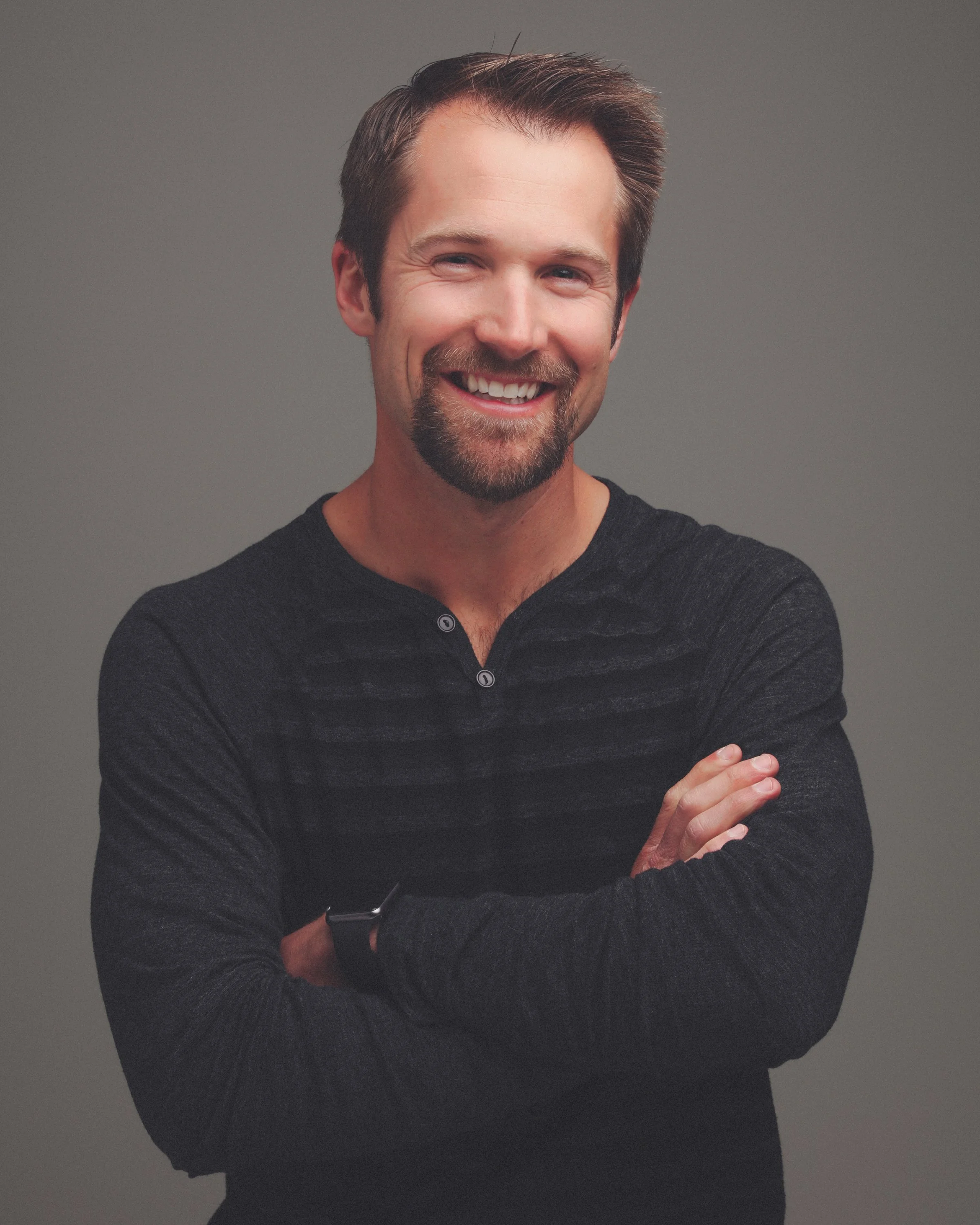 A smiling man with brown hair and a beard, wearing a dark long-sleeve shirt, standing with arms crossed against a gray background.
