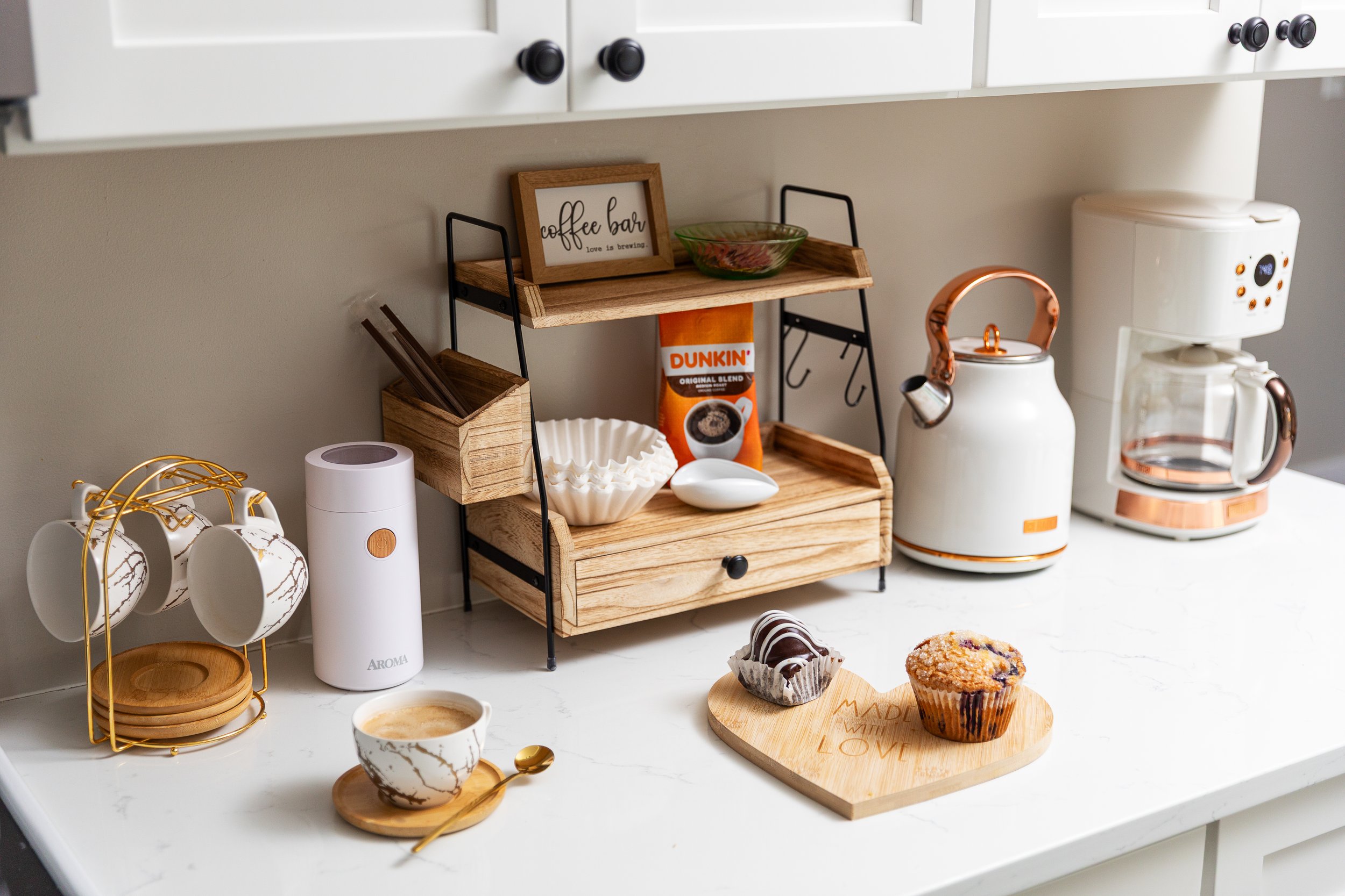 A kitchen countertop with coffee-making items, including a coffee grinder, a teapot, a coffee maker, and a wooden tray with muffins, cups, and a coffee box, in a modern and stylish setting.