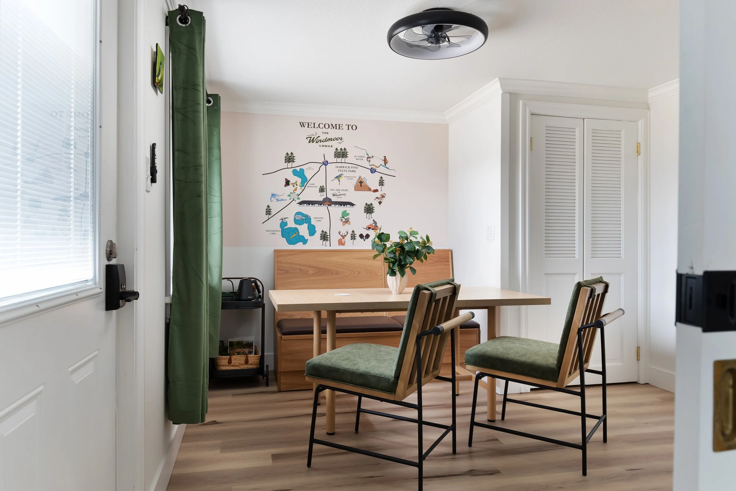 Dining area with a wooden table, green cushioned chairs, a small wooden bench, a potted plant, a wall map of Windmoor Lodge, and a ceiling light fixture.