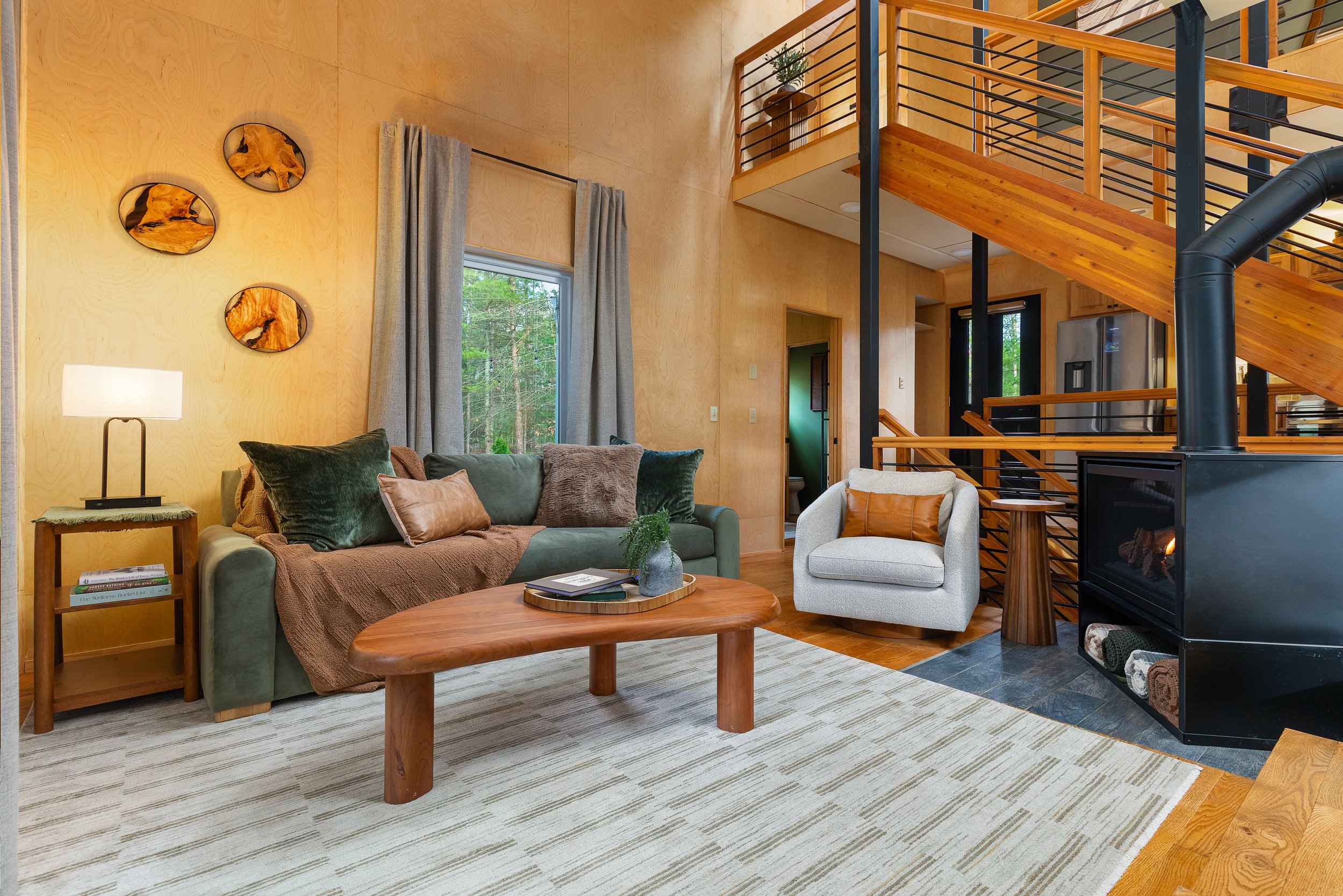 Living room with wooden walls, green sofa with cushions, white armchair, wood coffee table, fireplace, stairs made of wood and black metal, window with curtains, and decorative wall art.