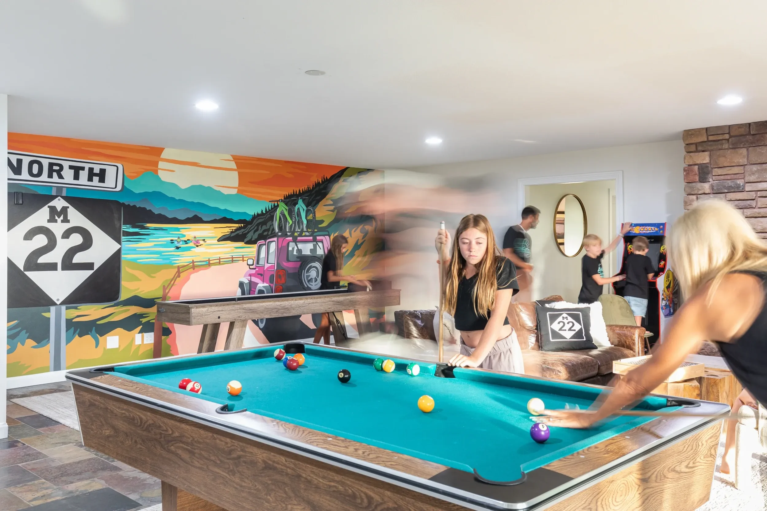 Kids playing pool in a game room with a colorful mountain and road sign mural on the wall.