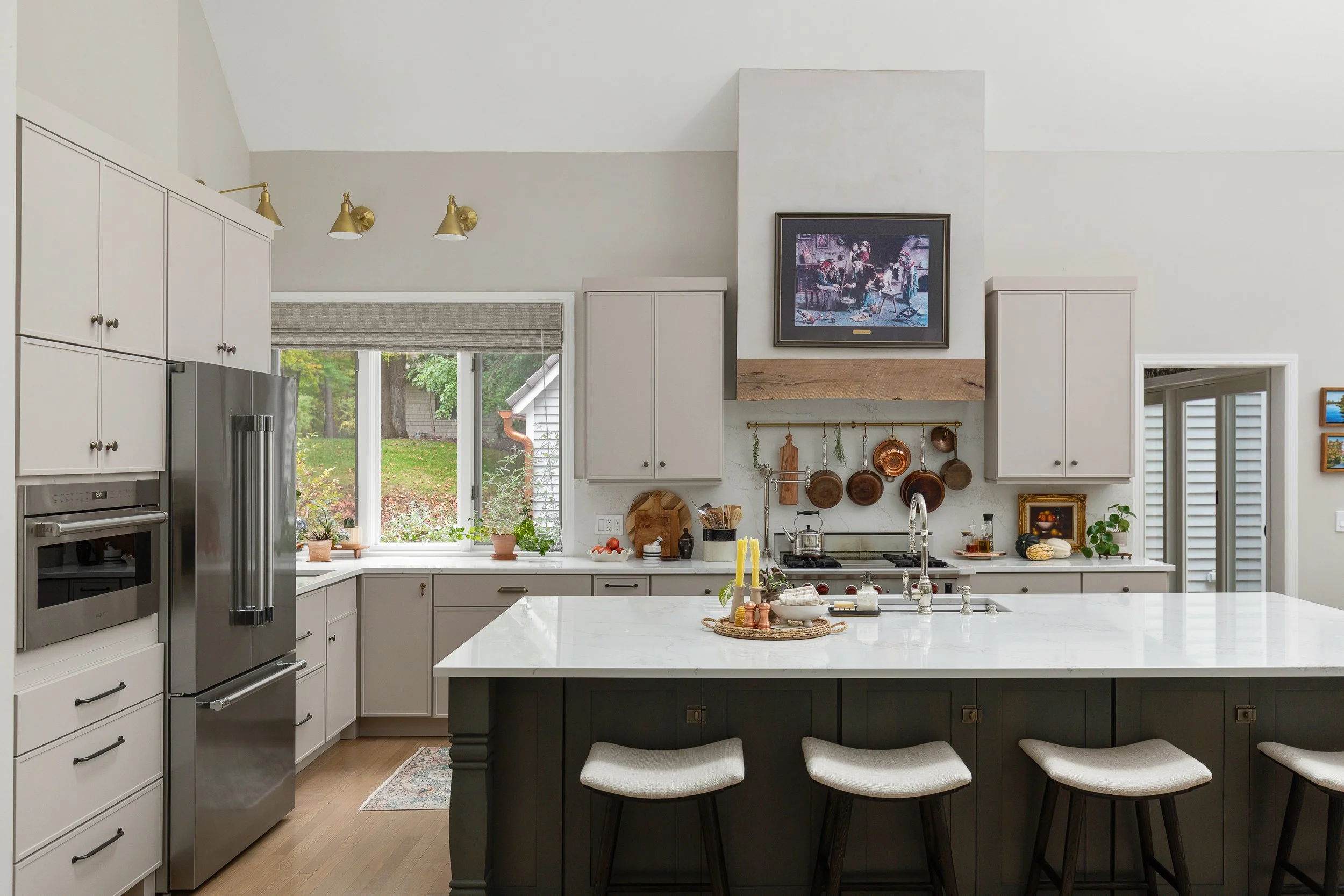 Modern kitchen with white cabinets, stainless steel refrigerator, and a large island with barstools. A window with plants overlooks a yard, and there are copper pots hanging above the stove. A TV is mounted above a wooden beam in the center.