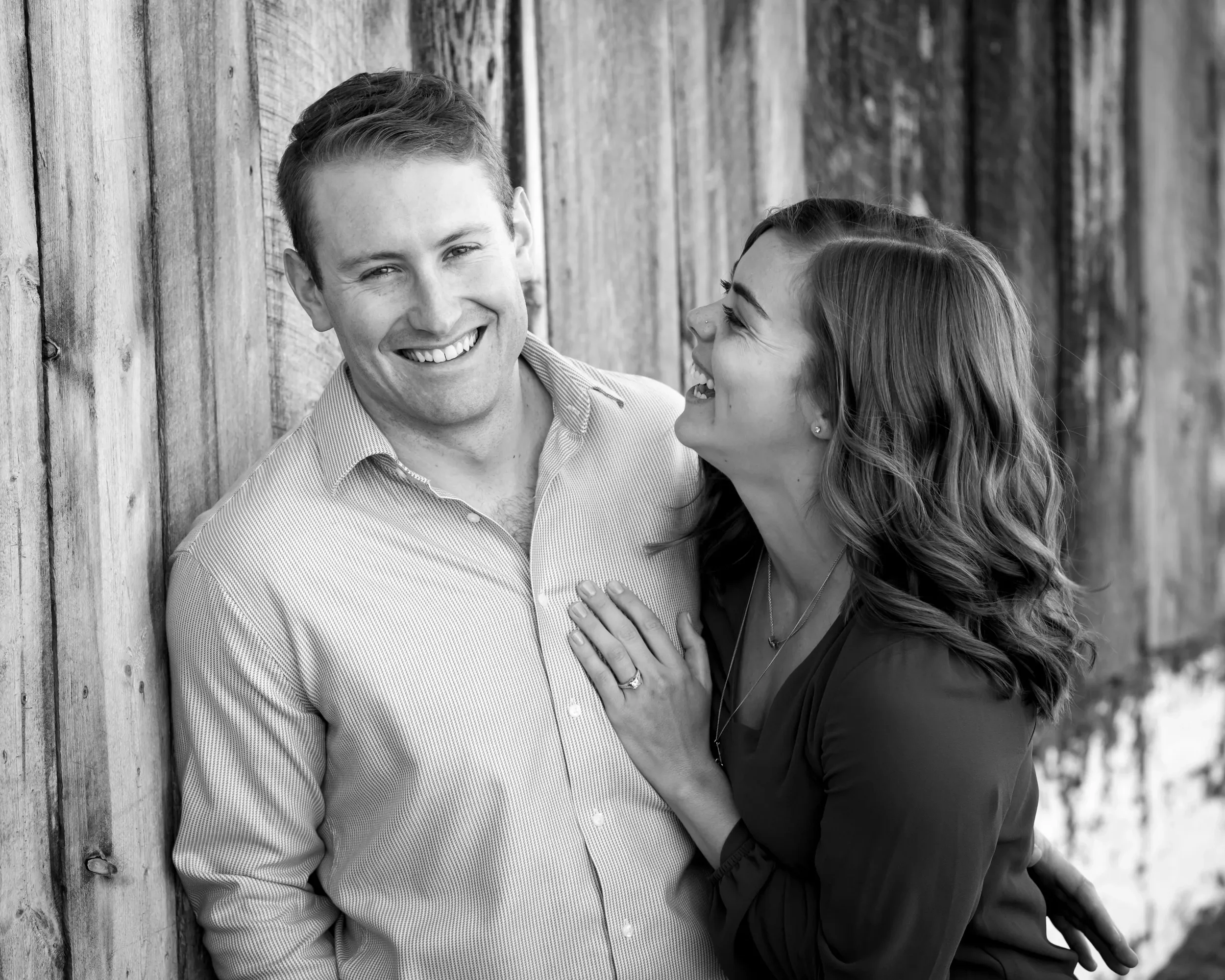 A happy couple leaning against a wooden wall, smiling and looking at each other, with the woman touching the man's chest.