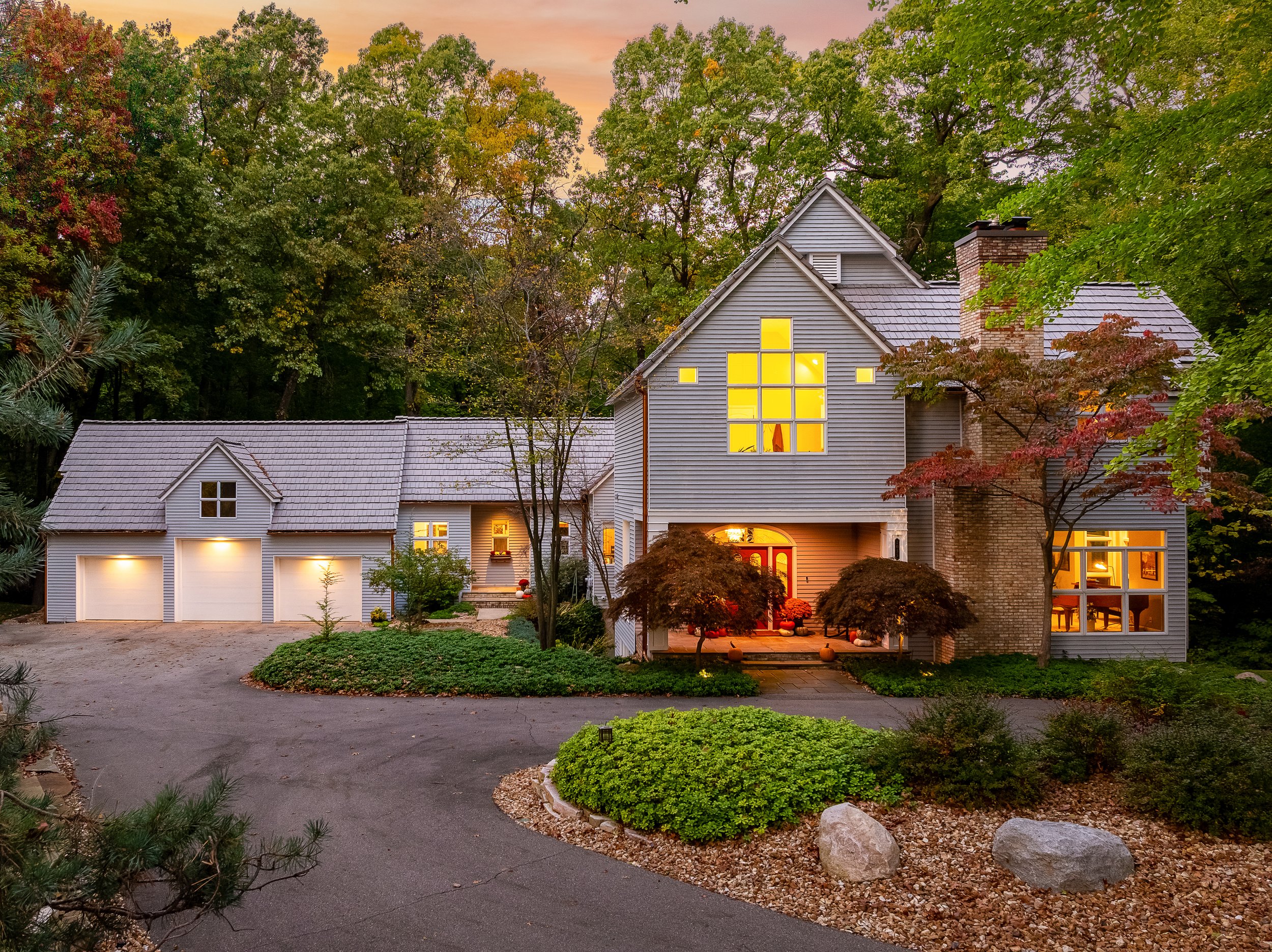 A large house with lit windows in a wooded area at sunset, featuring a driveway, front porch, and brick chimney surrounded by trees and landscaped bushes.