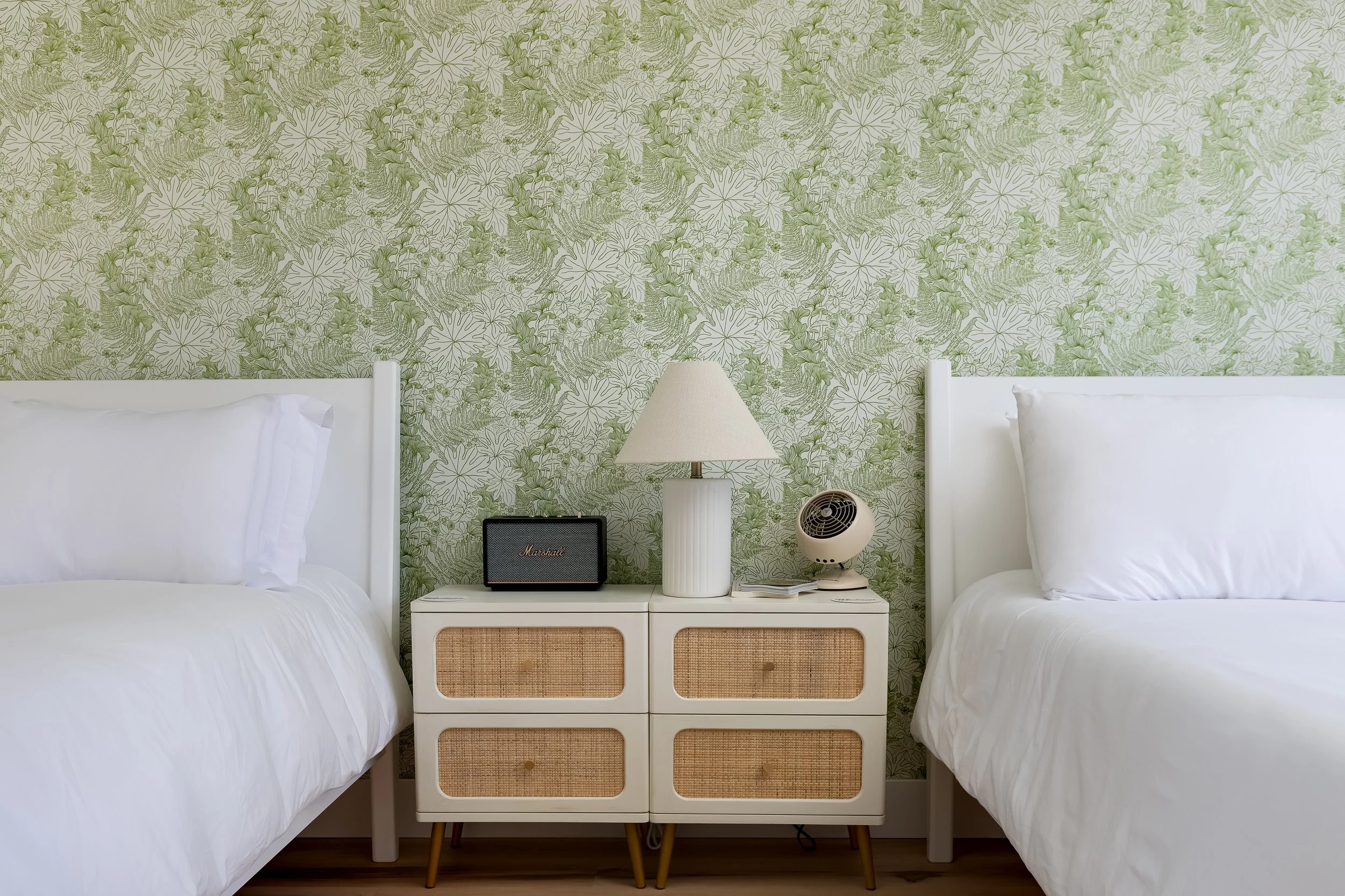 Two beds with white bedding and pillows separated by a white nightstand with a lamp, a small speaker, a vintage fan, and a phone, against a green floral wallpapered wall.