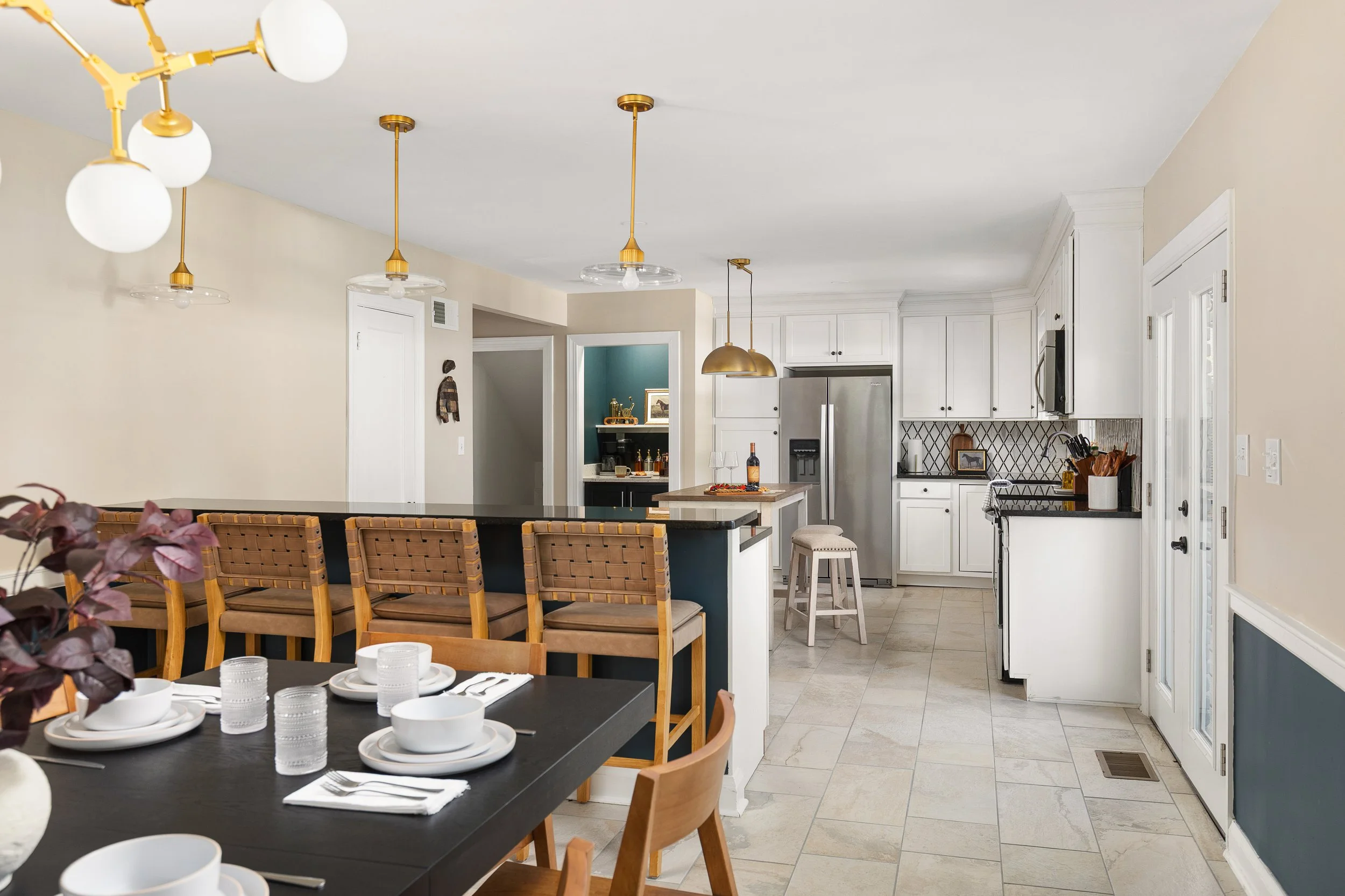 Modern kitchen and dining area with white cabinets, black countertops, a stainless steel refrigerator, and a black dining table set with white dishware and glasses, featuring a decorative flower centerpiece.