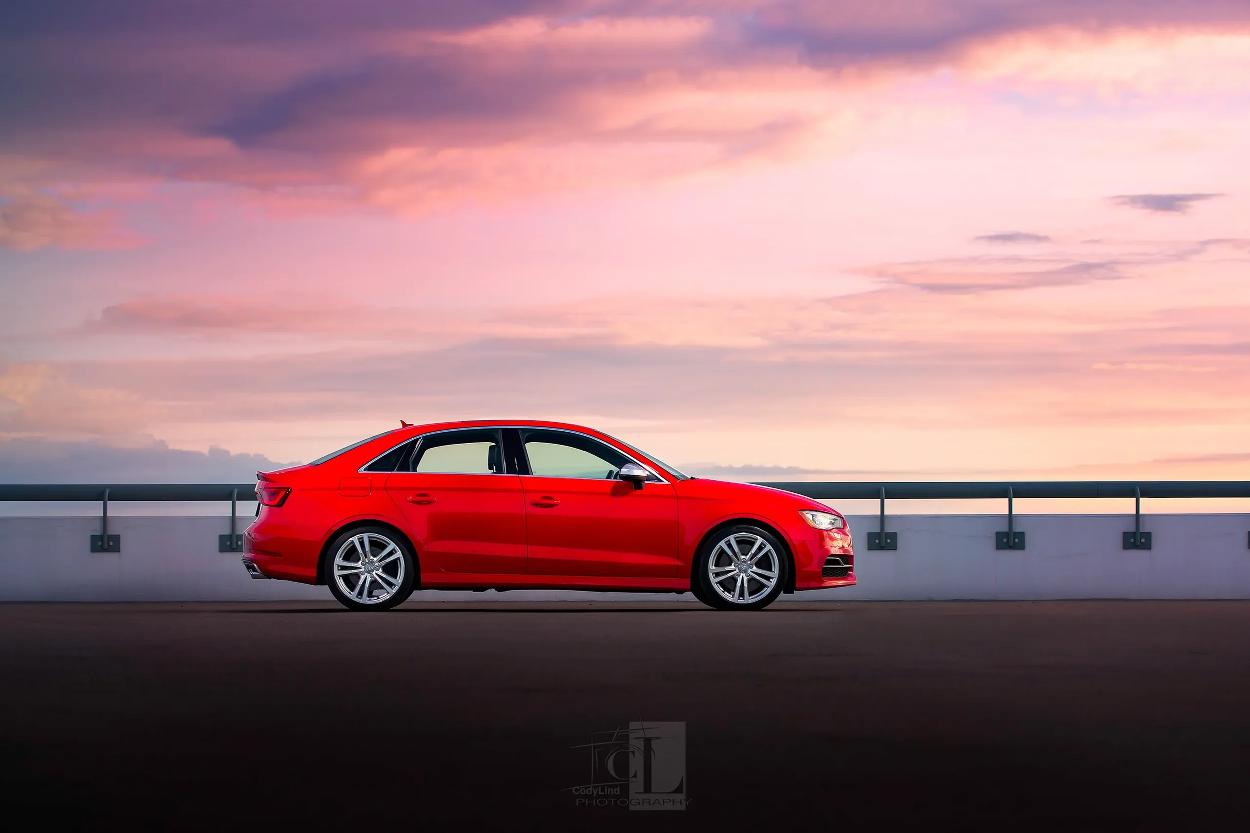 A red sedan car parked on an empty road during sunset or sunrise with colorful sky and clouds.