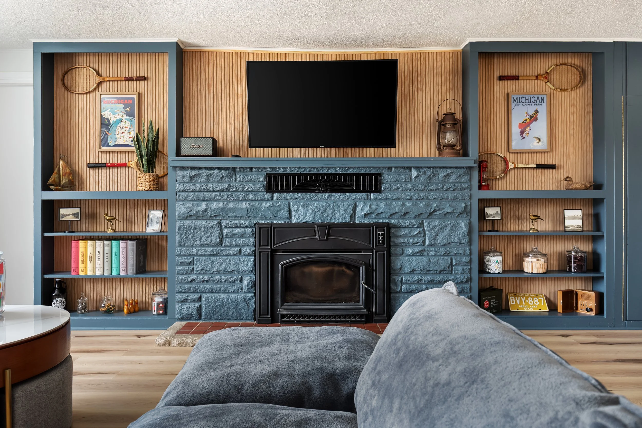 Living room with a blue brick fireplace, a flat-screen TV above, and built-in shelves on either side displaying vintage tennis rackets, framed artwork, small collectibles, and books. A gray sofa and a small side table are visible.