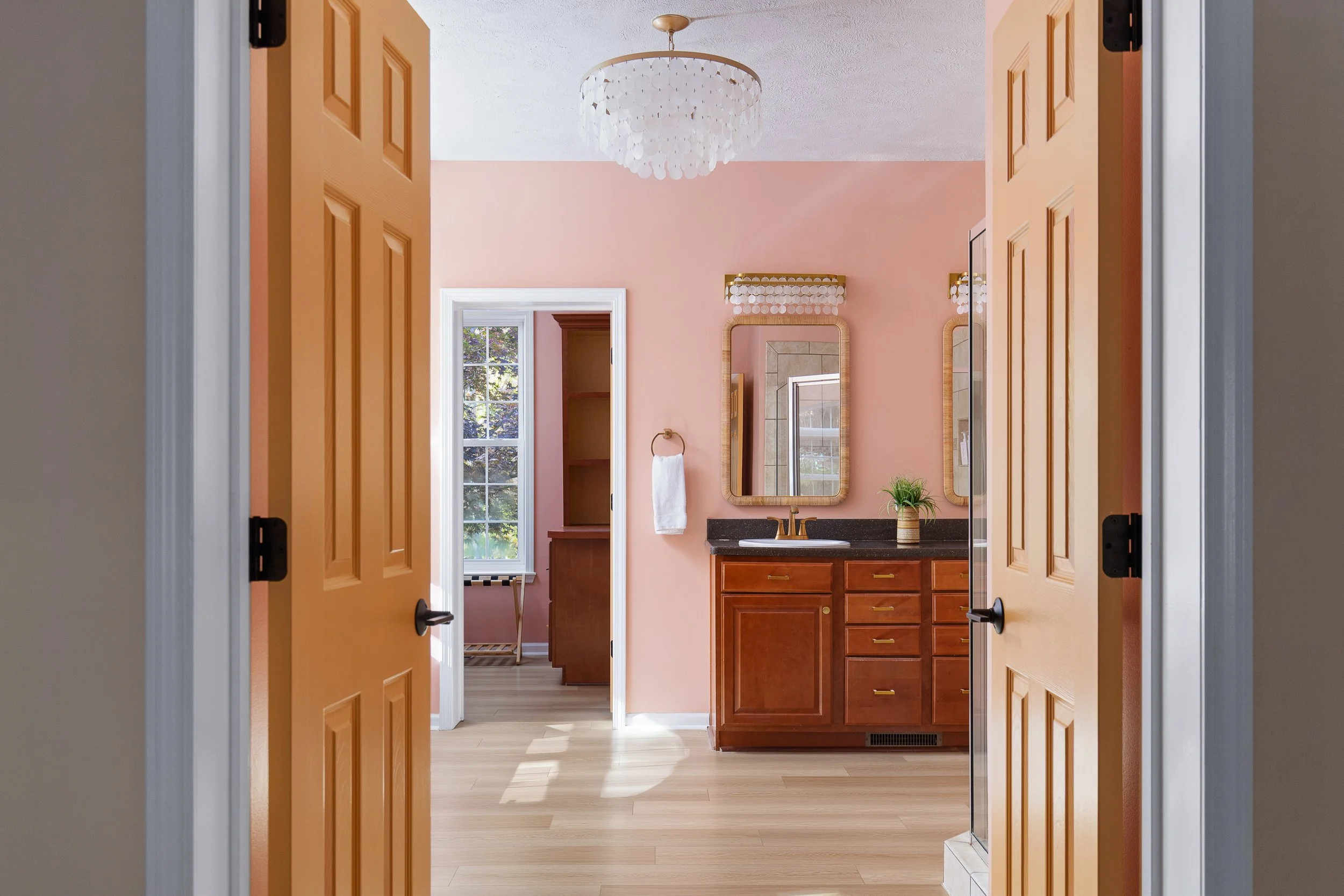 View through double wooden doors into a bathroom with pink walls, a black countertop, a wooden vanity, and a mirror, with a window revealing trees outside.