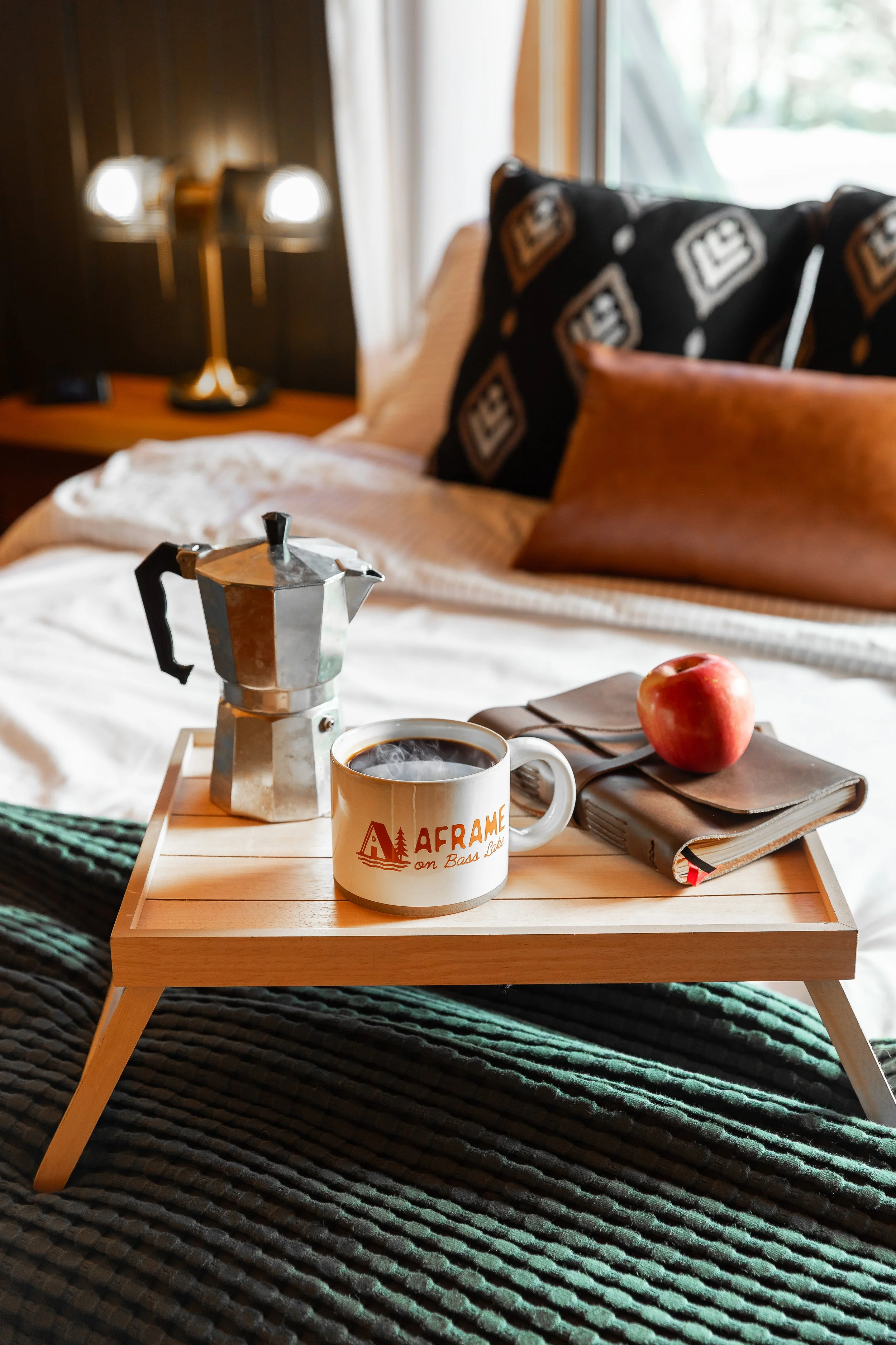 A tray on a bed, holding a steaming cup of coffee, a moka pot, an apple, a leather journal, and a pen, with a cozy bedroom in the background.