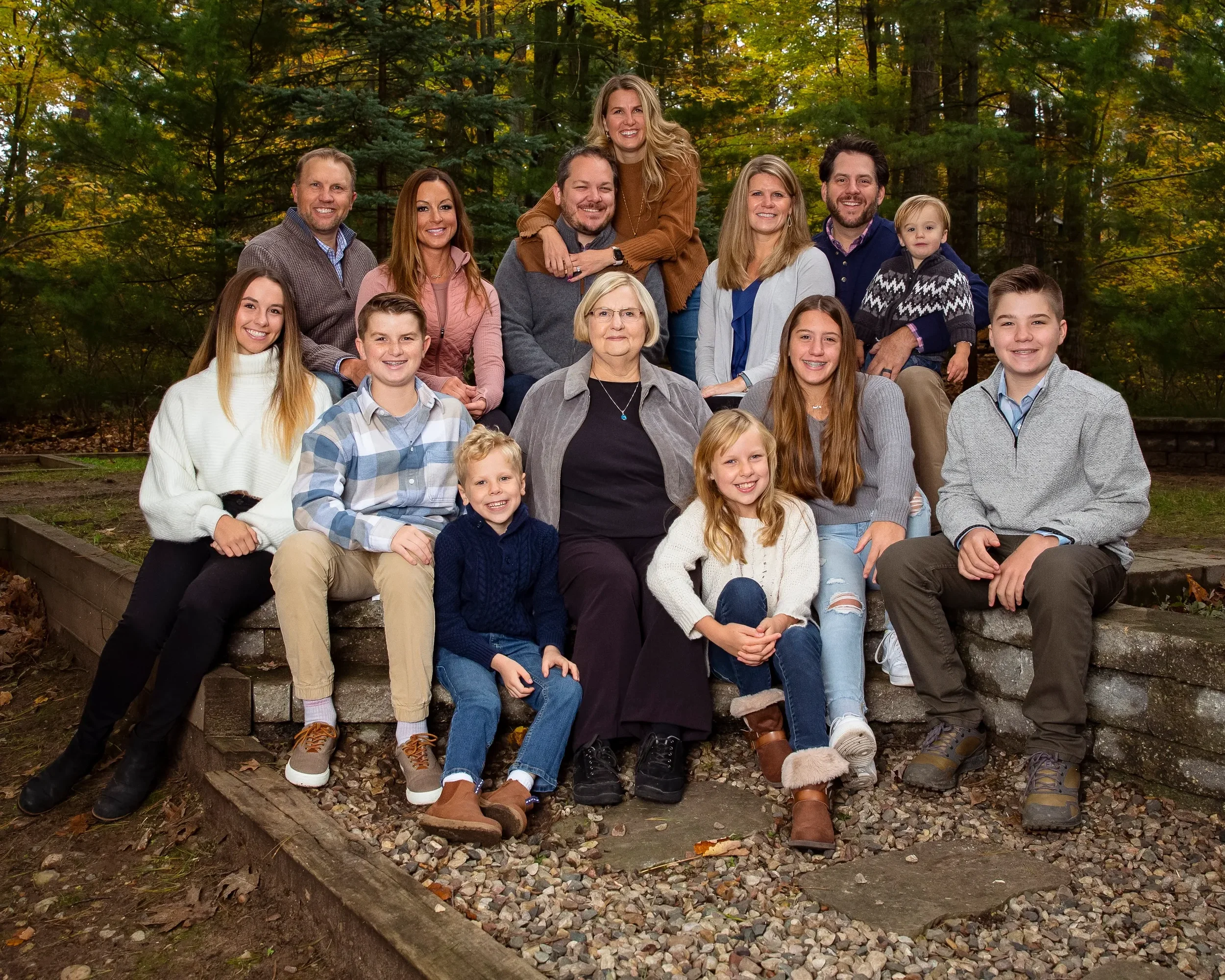 A large family photo outdoors in autumn, with 16 members including children, teenagers, adults, and an elderly woman, surrounded by trees with fall foliage.