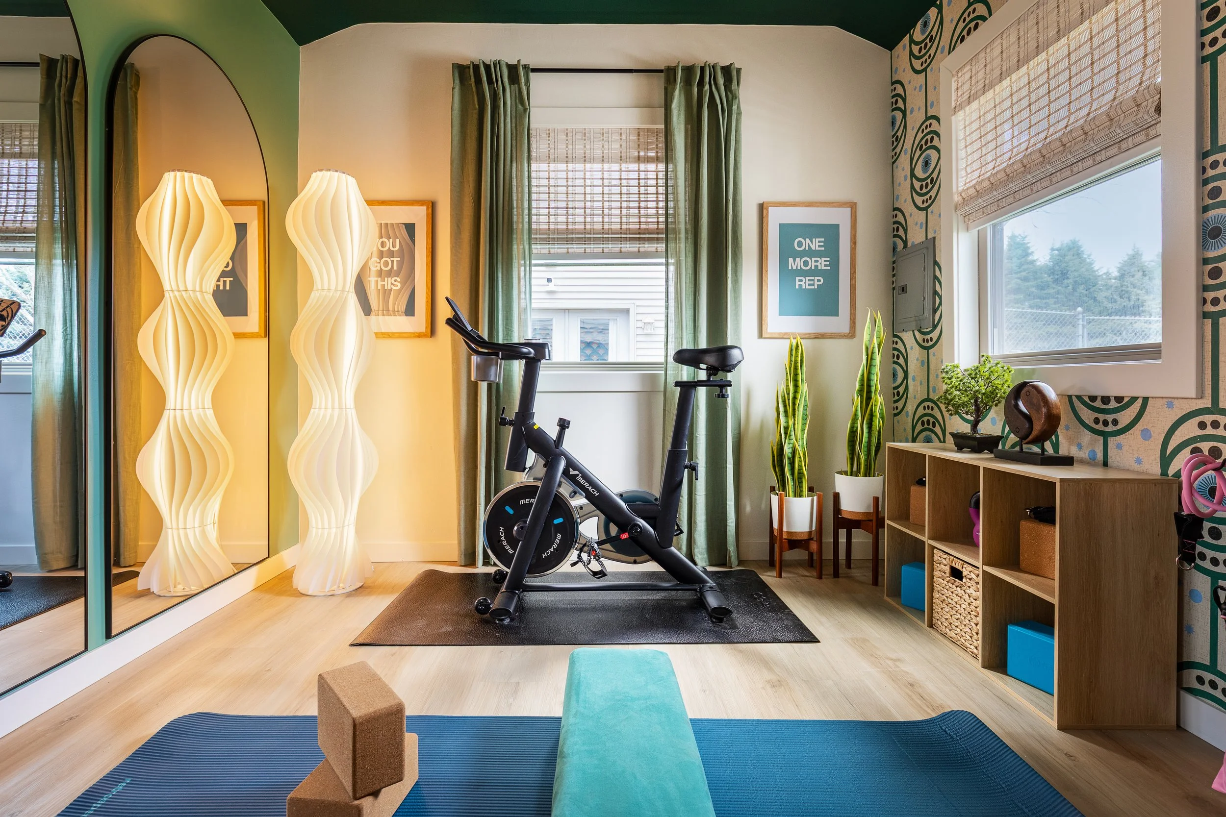 Home gym with exercise bike, yoga mat, foam blocks, decorative lamps, plants, shelving unit, and framed art, with windows and green curtains.