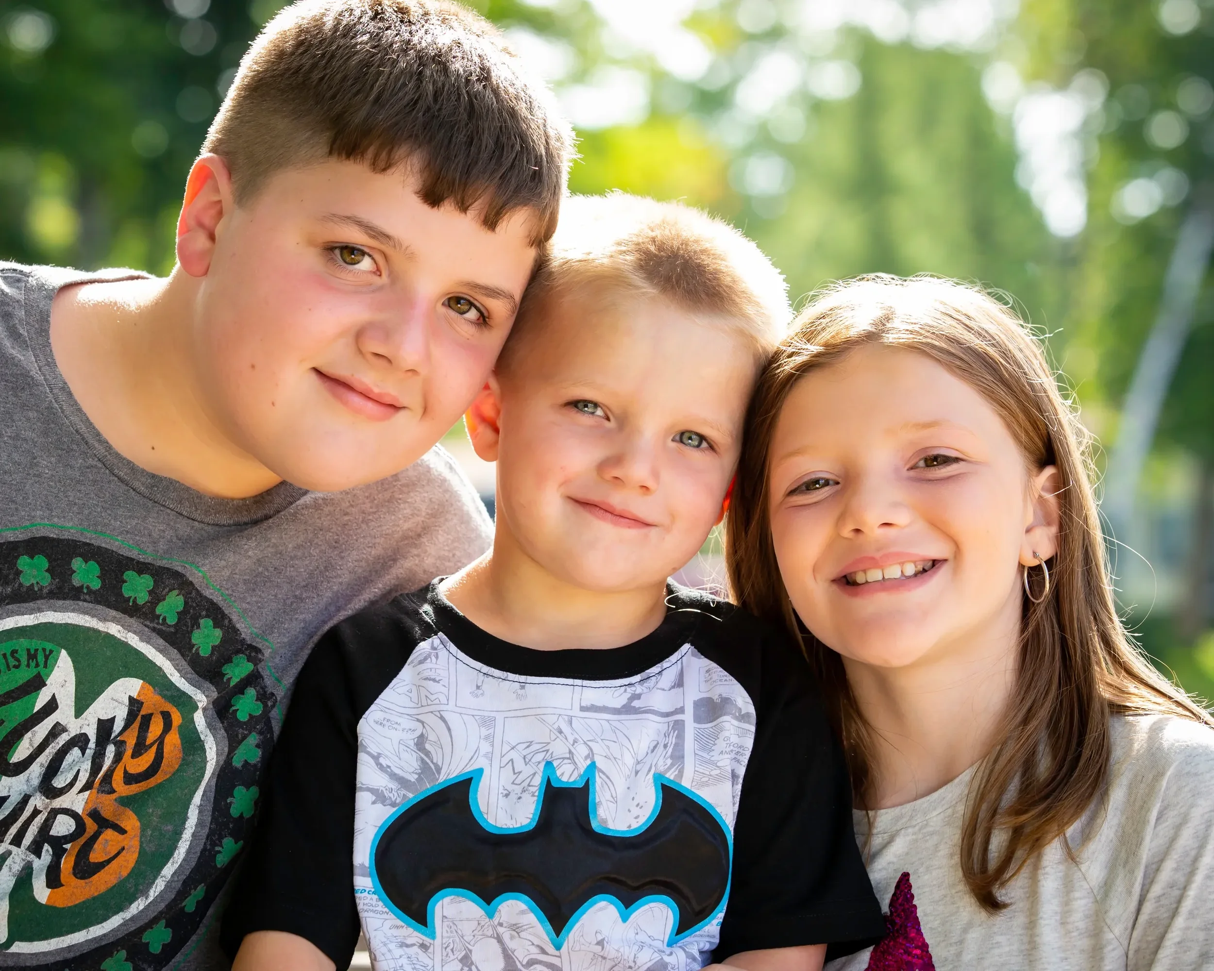 Three children smiling and posing outdoors on a sunny day.