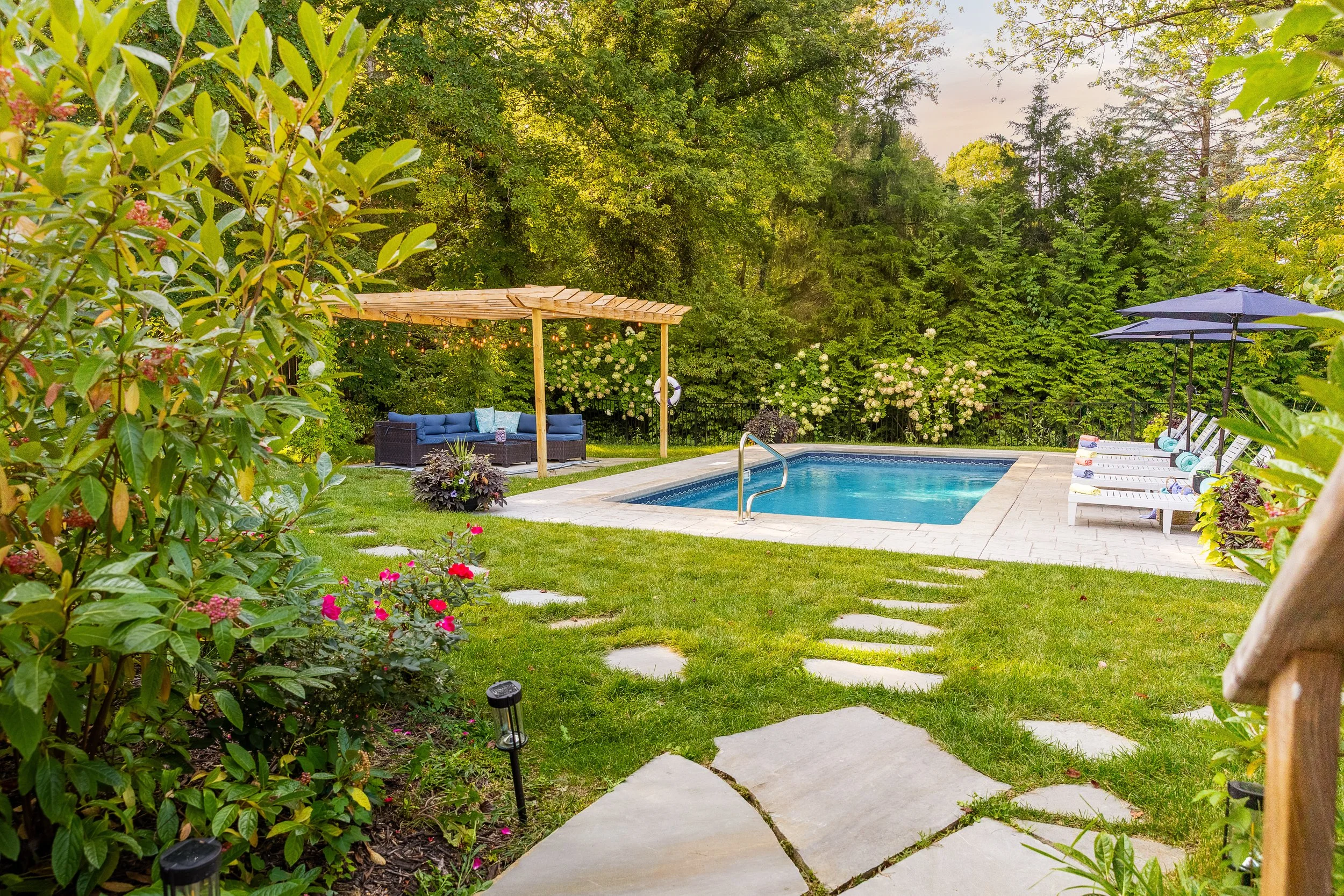 A backyard with a swimming pool, surrounded by green grass and trees. There is a wooden pergola with outdoor seating, a pair of white lounge chairs with umbrellas, and a stone pathway leading to the pool.