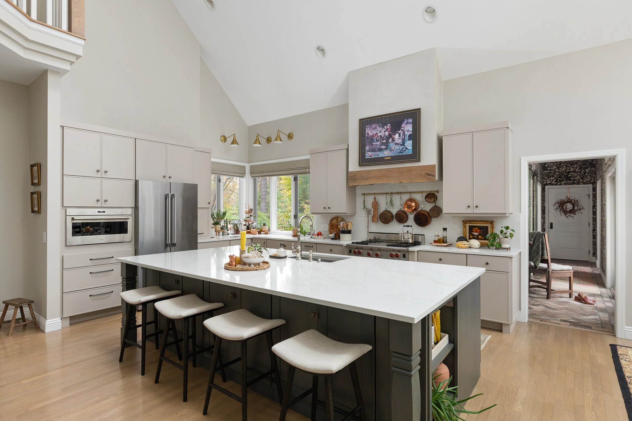 Modern kitchen with white cabinets, a large center island with a white marble top, and stainless steel appliances. Copper pots hang above the stove, and a window provides natural light. There's a doorway to another room decorated with a wreath hanging on the door.