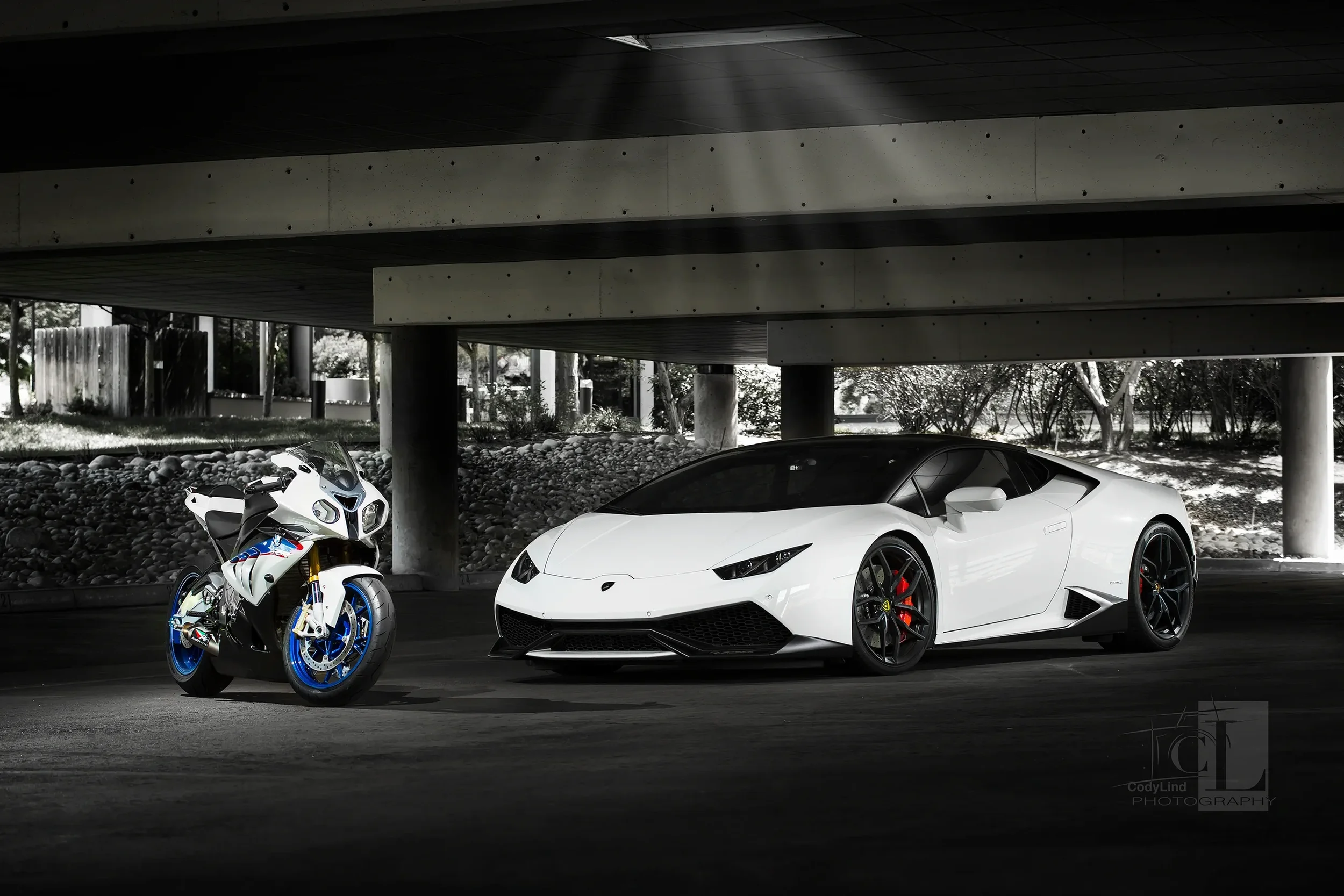 A white Lamborghini sports car and a white and blue Ducati motorcycle parked side by side in a covered parking area with concrete structure and trees visible in the background.