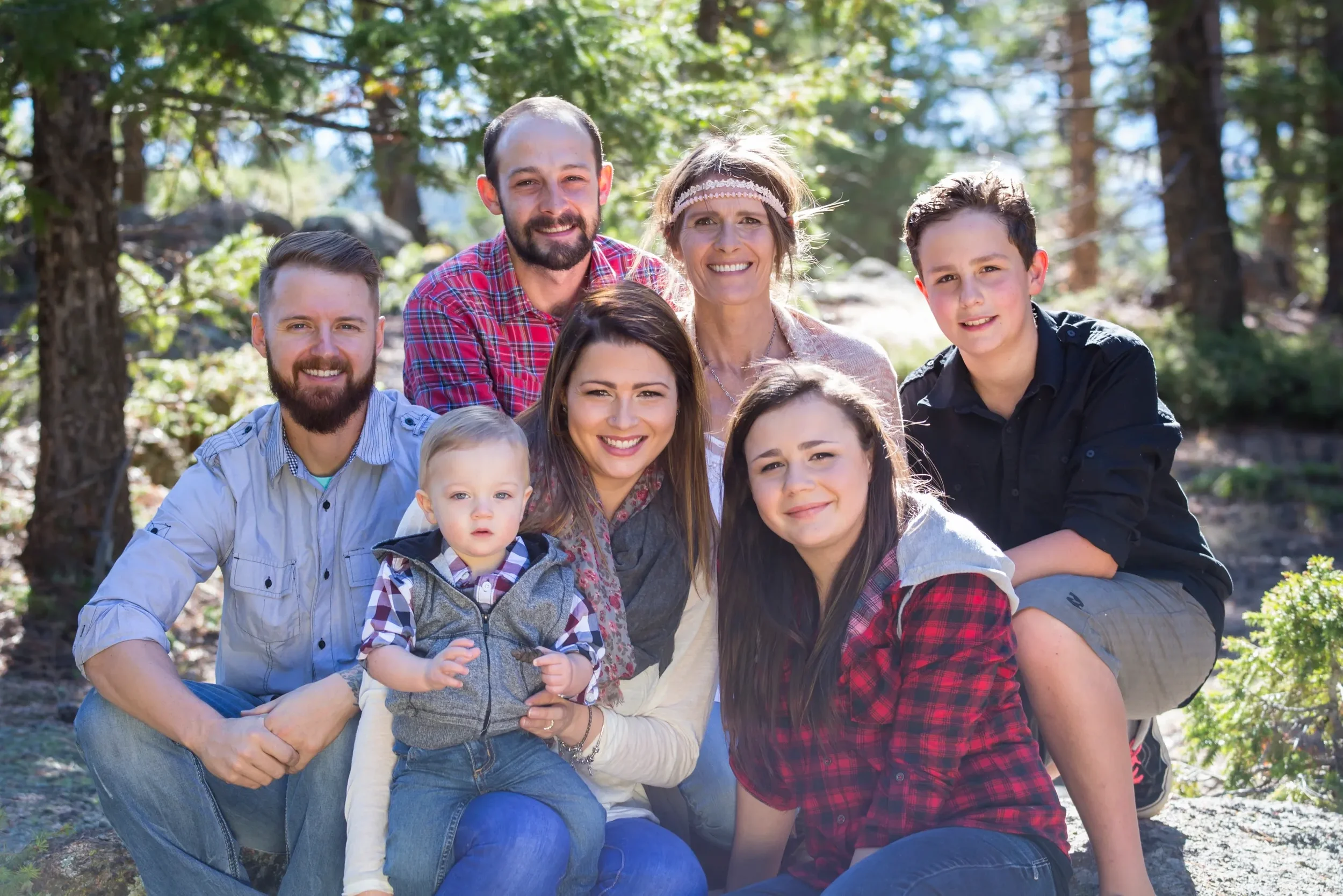 A family of eight posing outdoors in a forested area with sunlight filtering through trees, smiling at the camera.