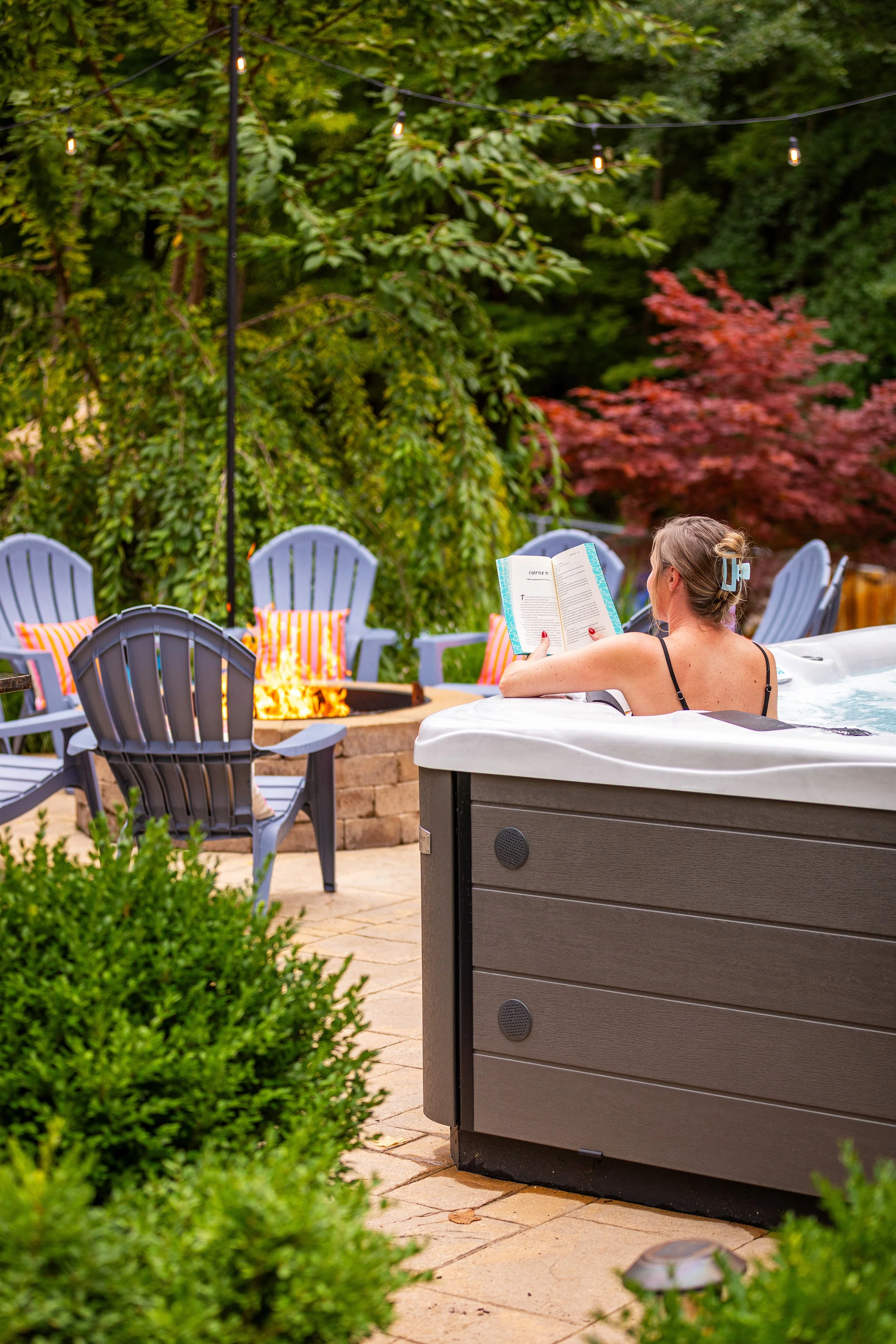 A woman relaxing in a hot tub outdoors, reading a book, with string lights above, surrounded by colorful chairs, a fire pit, and lush greenery.