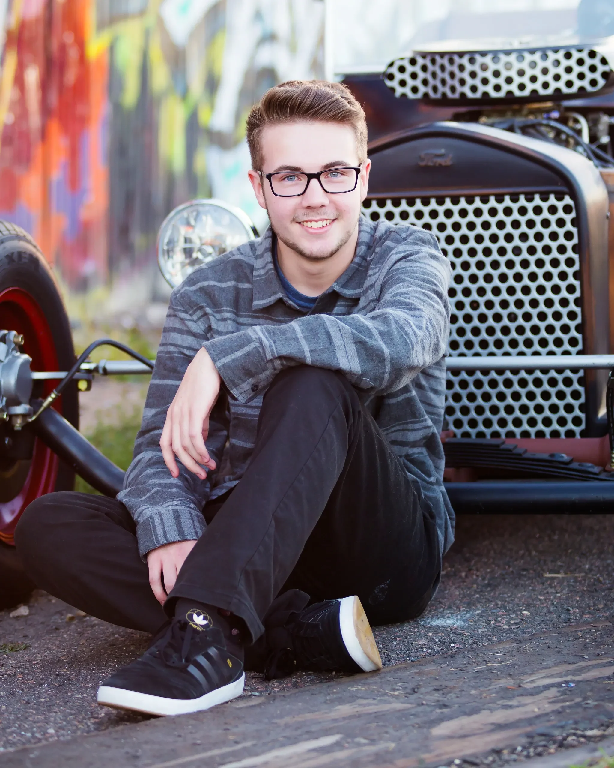 A young man with glasses, wearing a gray plaid shirt and black pants, sitting on the ground in front of a vintage car with an open hood.