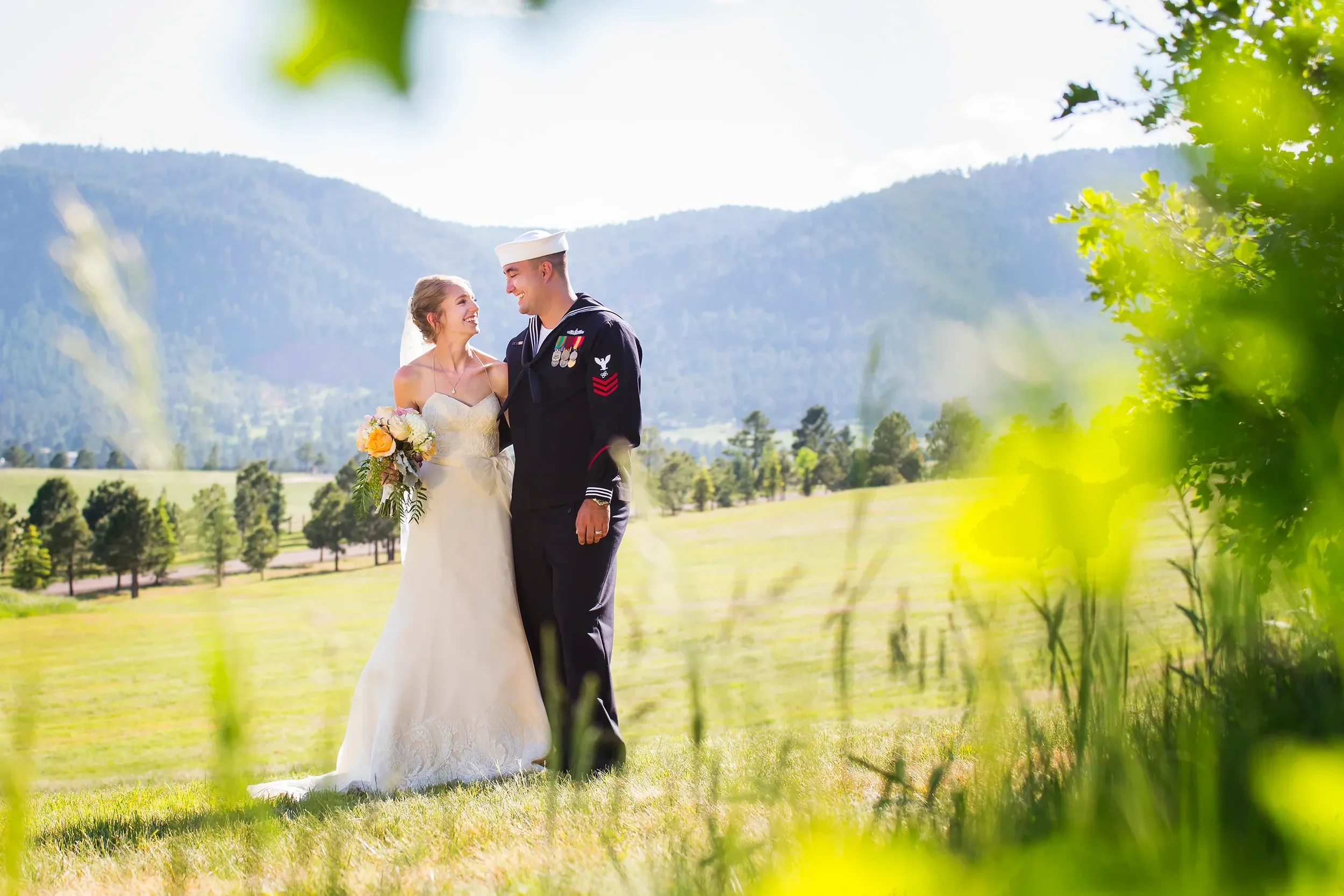 A bride and groom in wedding attire standing outdoors in a green field with mountains in the background, smiling at each other.